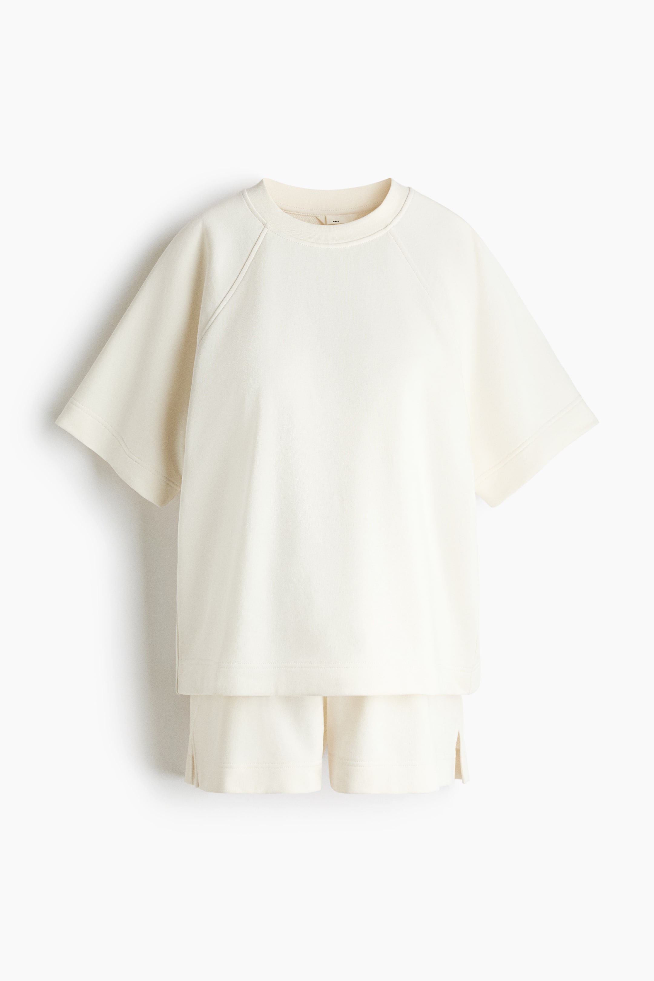 View larger image: A cream-colored set includes a t-shirt and shorts made from lightweight sweatshirt fabric, shown from the front. The t-shirt features a crew neck, raglan short sleeves, and a straight hem with side slits. The shorts display a gathered elastic waistband and small slits at the hems.