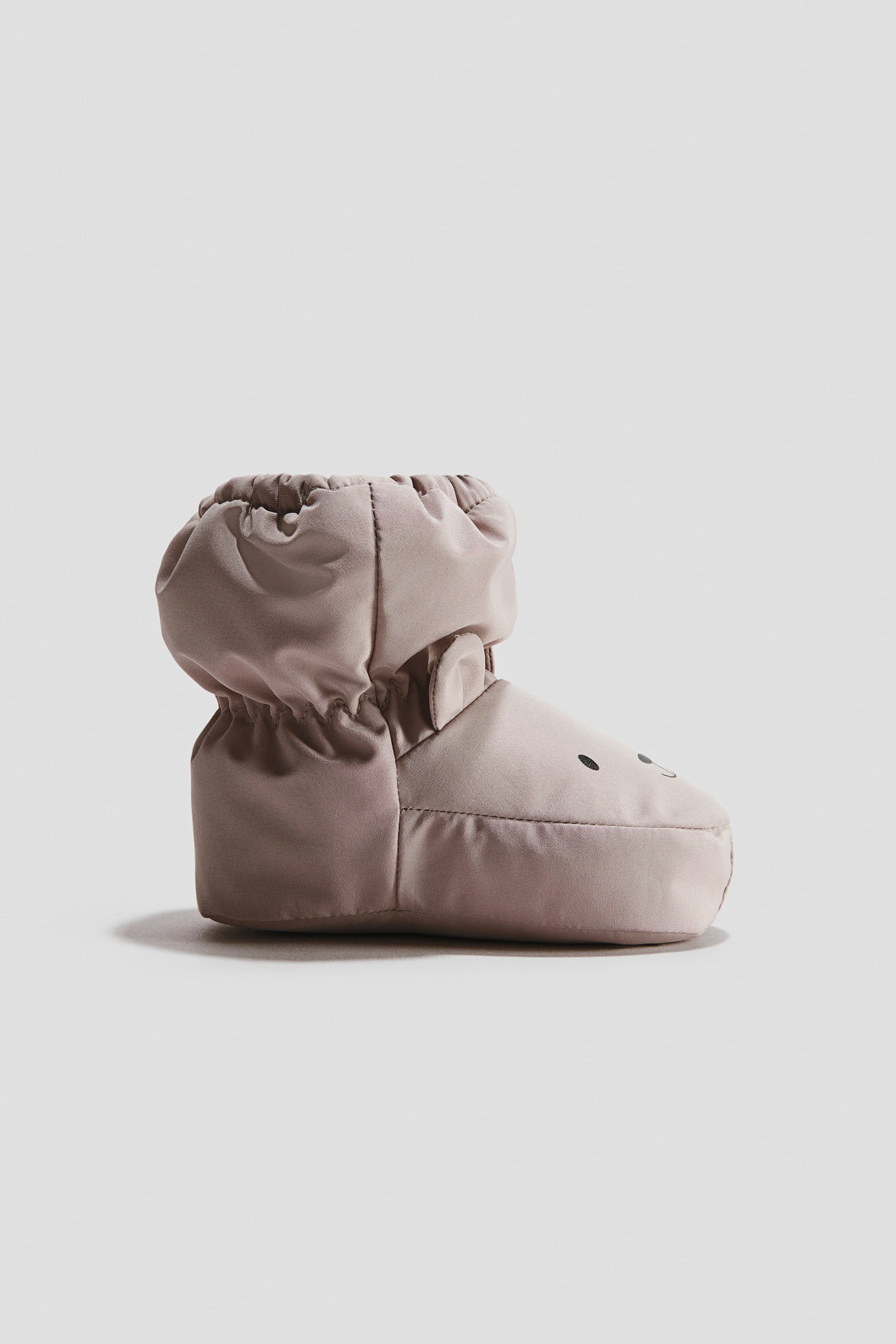 View larger image: A single light brown padded bootie is shown in side view, featuring a gathered ankle opening and small rounded ears. The toe area displays two black dots and a curved line, forming a simple face.