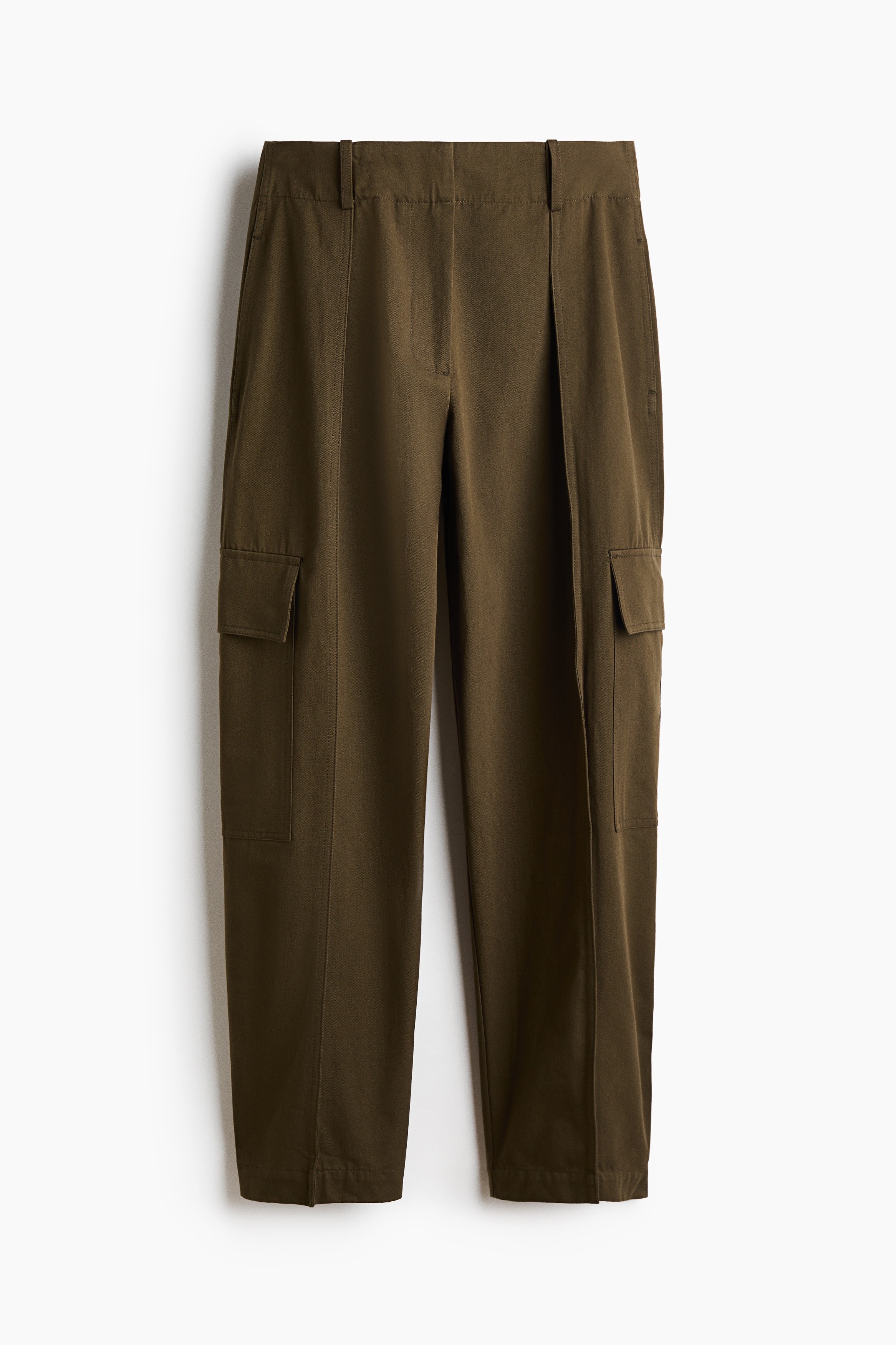 Olive green cotton trousers are shown from the front in a high-waisted, barrel-leg fit. The trousers feature prominent front pleats, visible centre seams running down each leg, and side flap pockets, styled with a loose white shirt that is partly tucked in to reveal the waistband.