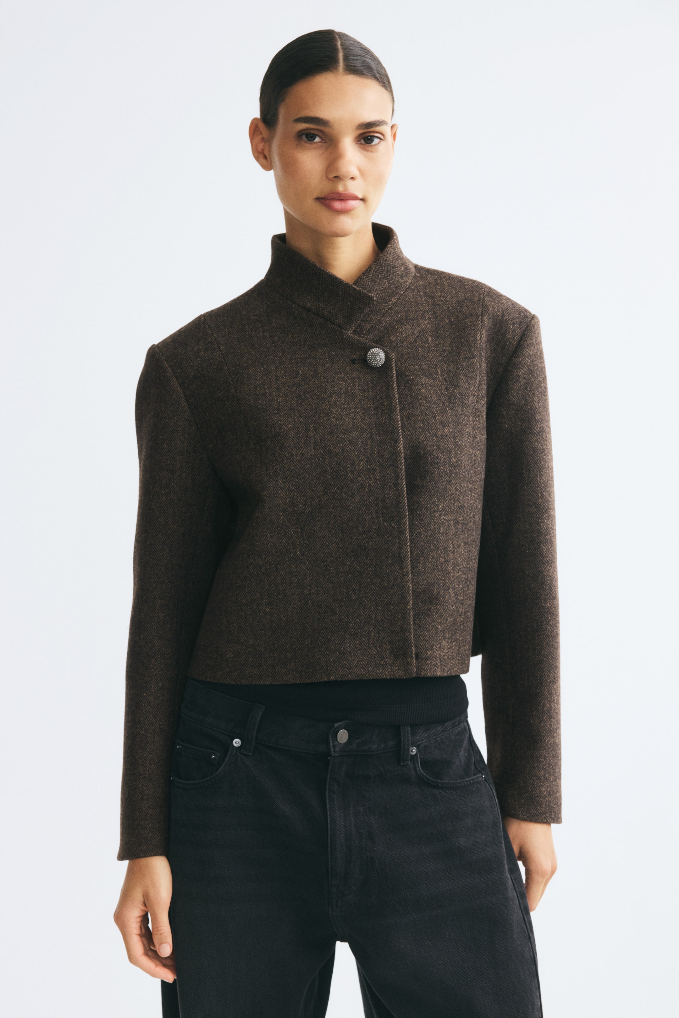 Agrandir l'image: Un blazer court en tweed brun à motif chevrons est porté par le mannequin, avec un petit col montant fermé par un seul bouton orné de strass. Ce blazer structuré et court est porté sur un haut noir et associé à un jean en denim noir.