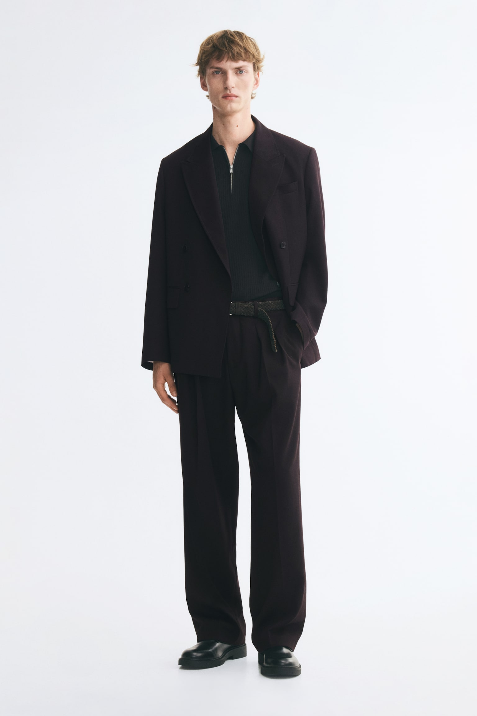 Suit trousers in a dark plum woven fabric are being worn by the model, who is standing with one hand resting inside a diagonal side pocket. The loose-fit trousers, featuring front pleats and straight legs with sharp creases, are styled with a woven belt, a matching double-breasted blazer, and black shoes. - 1