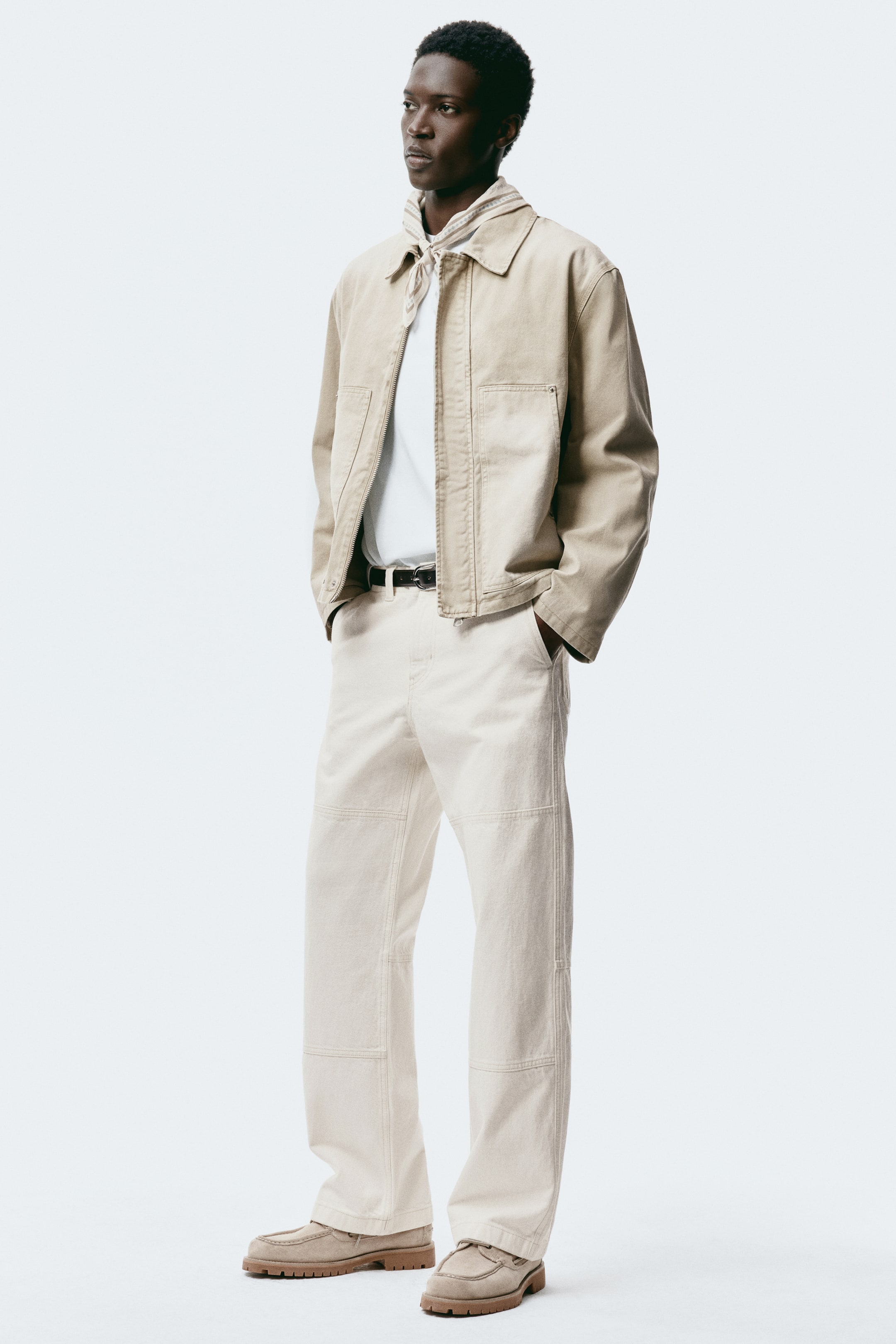 View larger image: A beige workwear-style jacket, featuring a collar, a full zipper, and two patch pockets, is layered over a white crew-neck T-shirt, with a light-coloured bandana tied around the neck. Light beige wide-leg pants complete the look, detailed with visible stitching and panelling, secured by a black belt, and paired with light-brown suede loafers with thick lug soles.