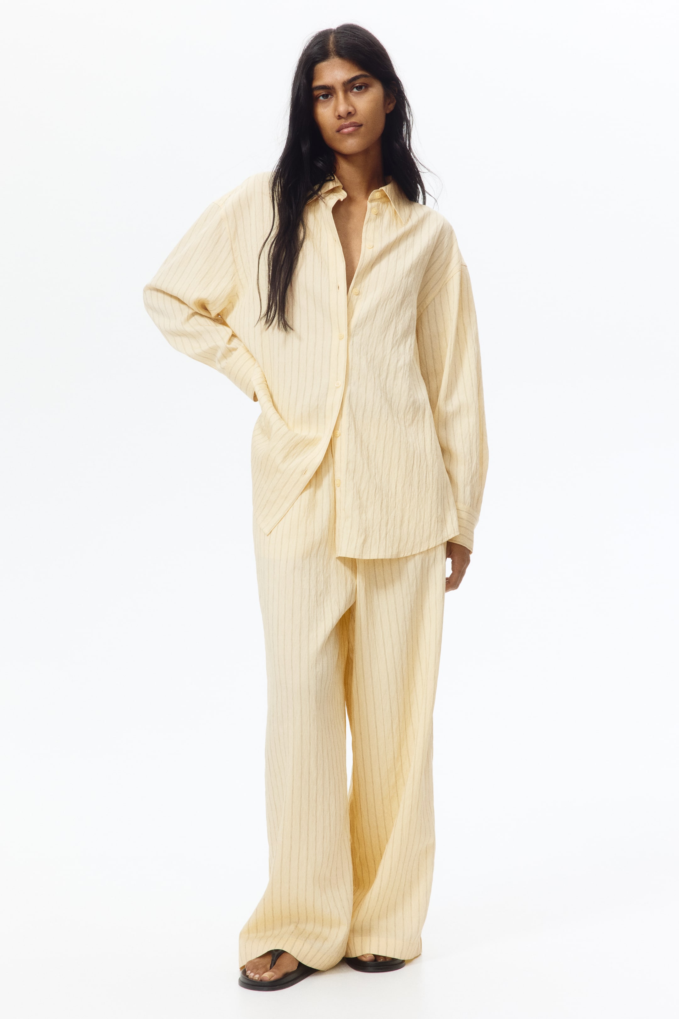 View larger image: A light yellow shirt and matching wide-leg trousers are showcased, facing forward. The fabric boasts a crinkled texture with subtle vertical pinstripes. The oversized shirt features a collar and long sleeves, with a few front buttons left undone.