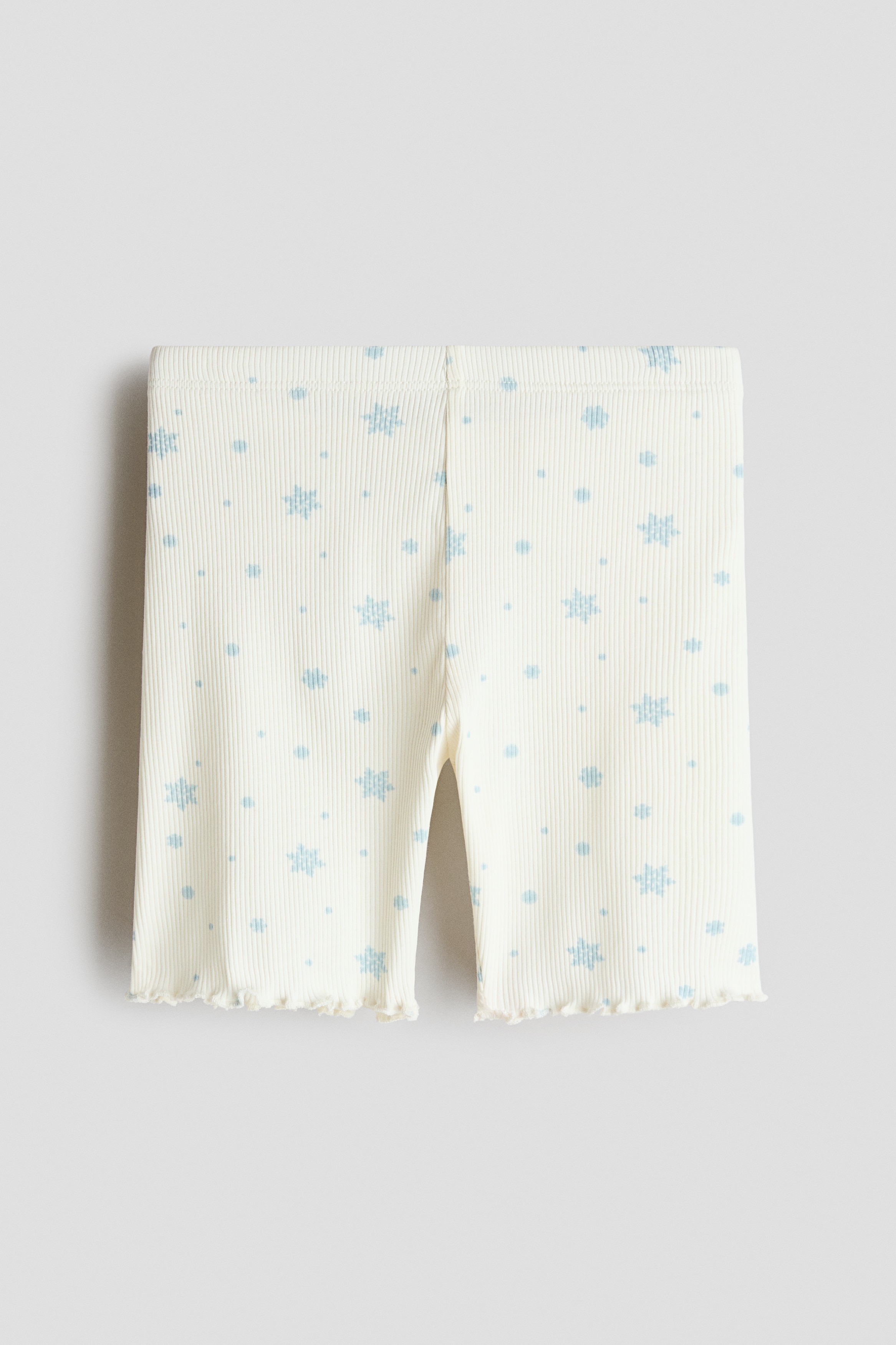 The model is wearing bike shorts in a soft, ribbed cream jersey with an all-over blue snowflake print and overlocked hems. The shorts are styled with a matching sleeveless top featuring a ruffled neckline.