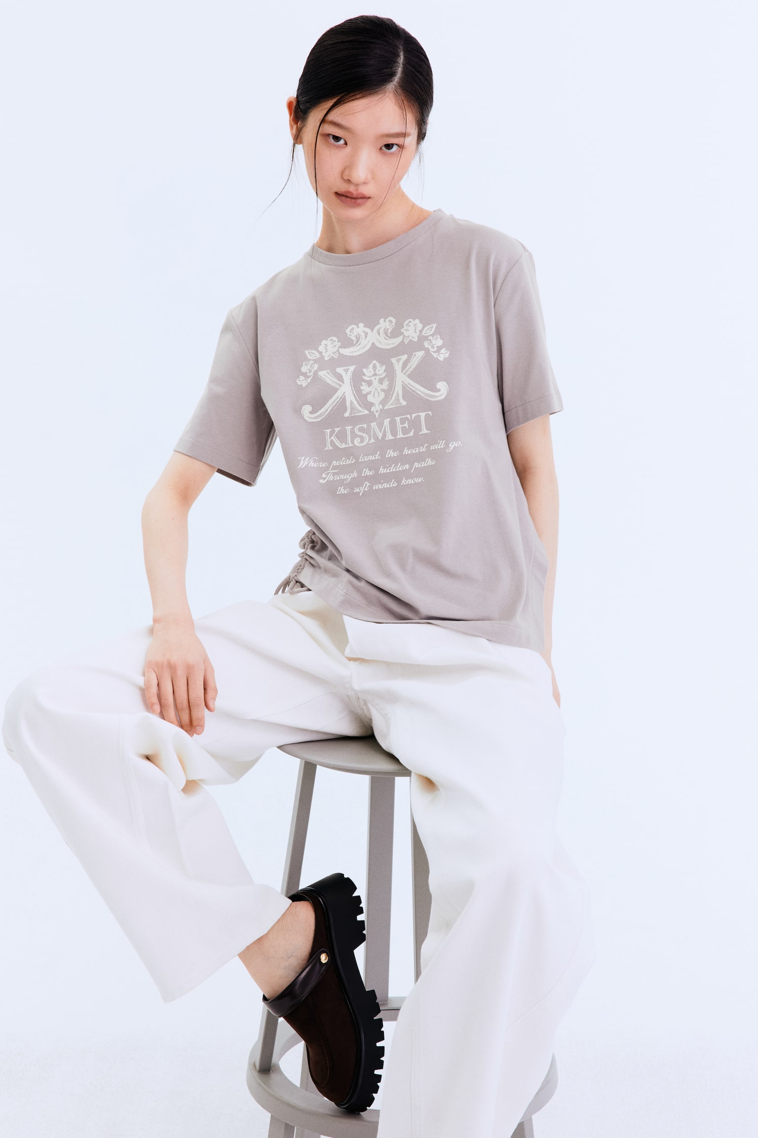 Presenting a taupe short-sleeved t-shirt with a crew neck, showcased in a forward-facing position. It features a white graphic print prominently centered on the front. The design includes two stylized 'K's encircled by floral motifs, the word 'KISMET', and an accompanying quote beneath. Additionally, a gathered detail is noticeable on the left side of the t-shirt. - 1