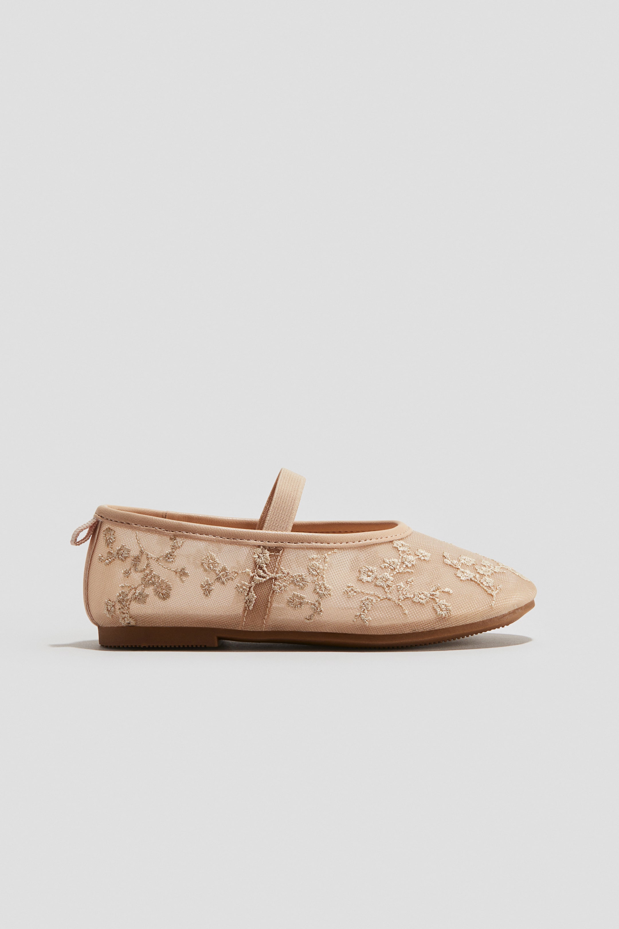 View larger image: A single light beige Mary Jane flat shoe is featuring a sheer mesh upper with matching floral embroidery, a single elasticized strap extending across the instep, a rounded toe, and a flat brown sole, presented in a side view.