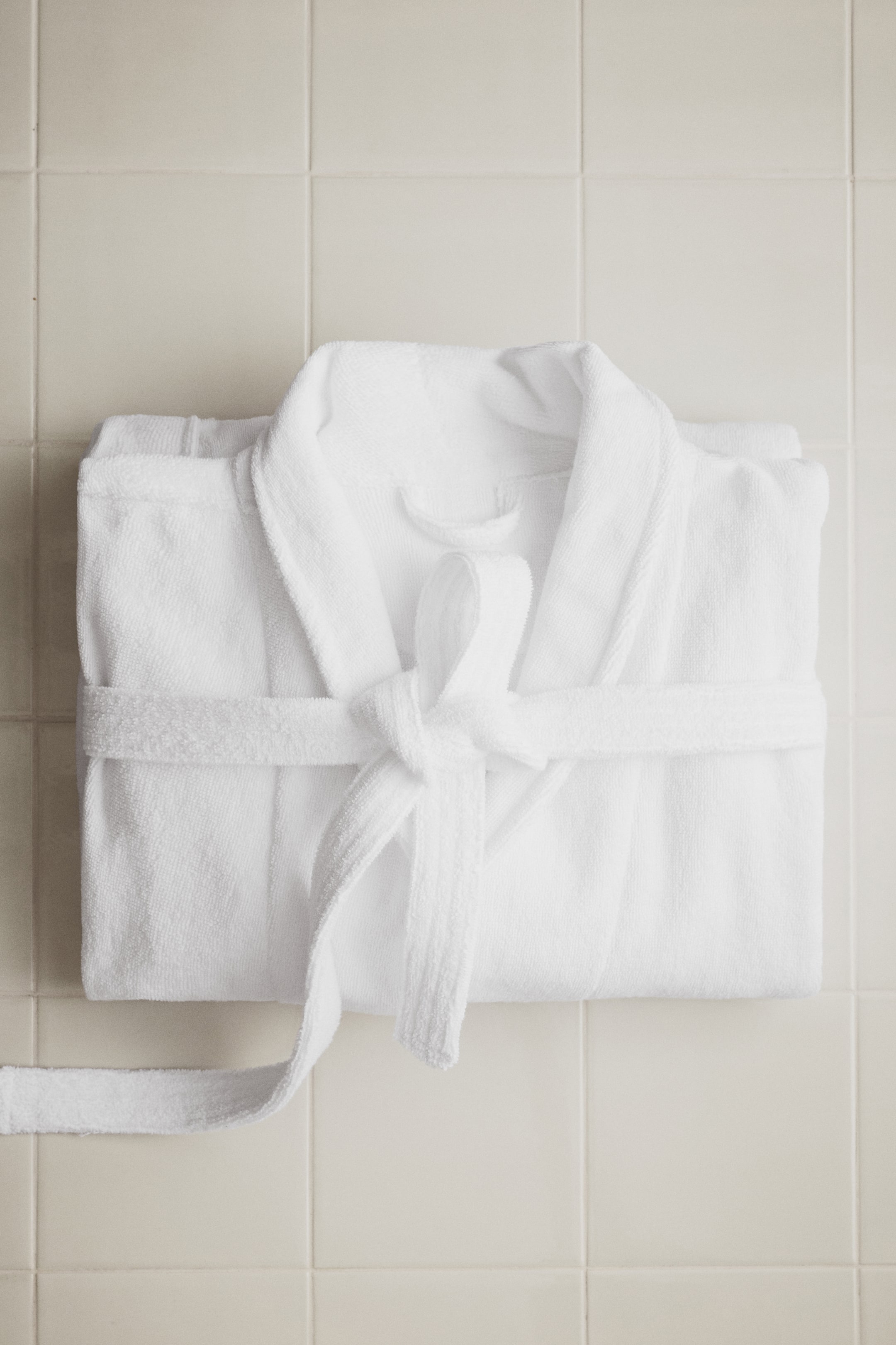 View larger image: A white terry dressing gown is lying folded, featuring a shawl collar, front pockets, and a detachable tie belt that is knotted at the waist.