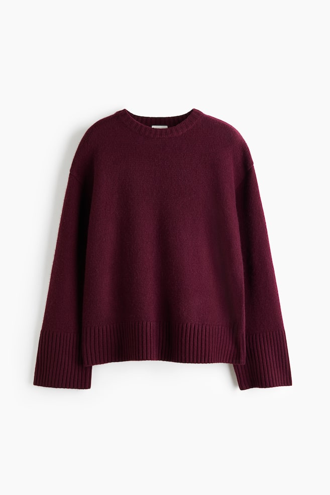 An oversized burgundy knit sweater is facing the front, showcasing dropped shoulders and long, wide sleeves. The sweater is featuring a ribbed crew neck, wide ribbed cuffs, and a ribbed hem. - 1