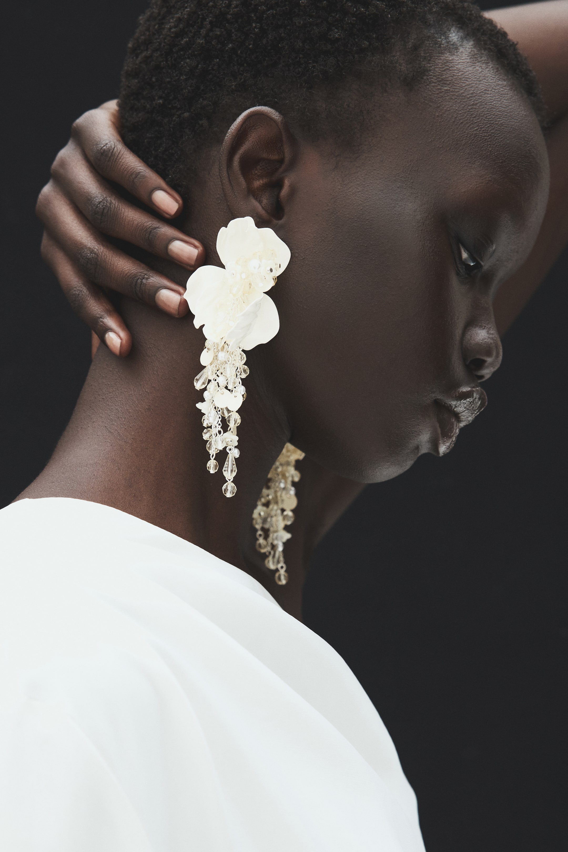 View larger image: Long, dangling earrings with white flower-shaped details and cascading strands of clear plastic beads are worn by the model. The earrings drape along the side of the neck as the model rests one hand behind their head.