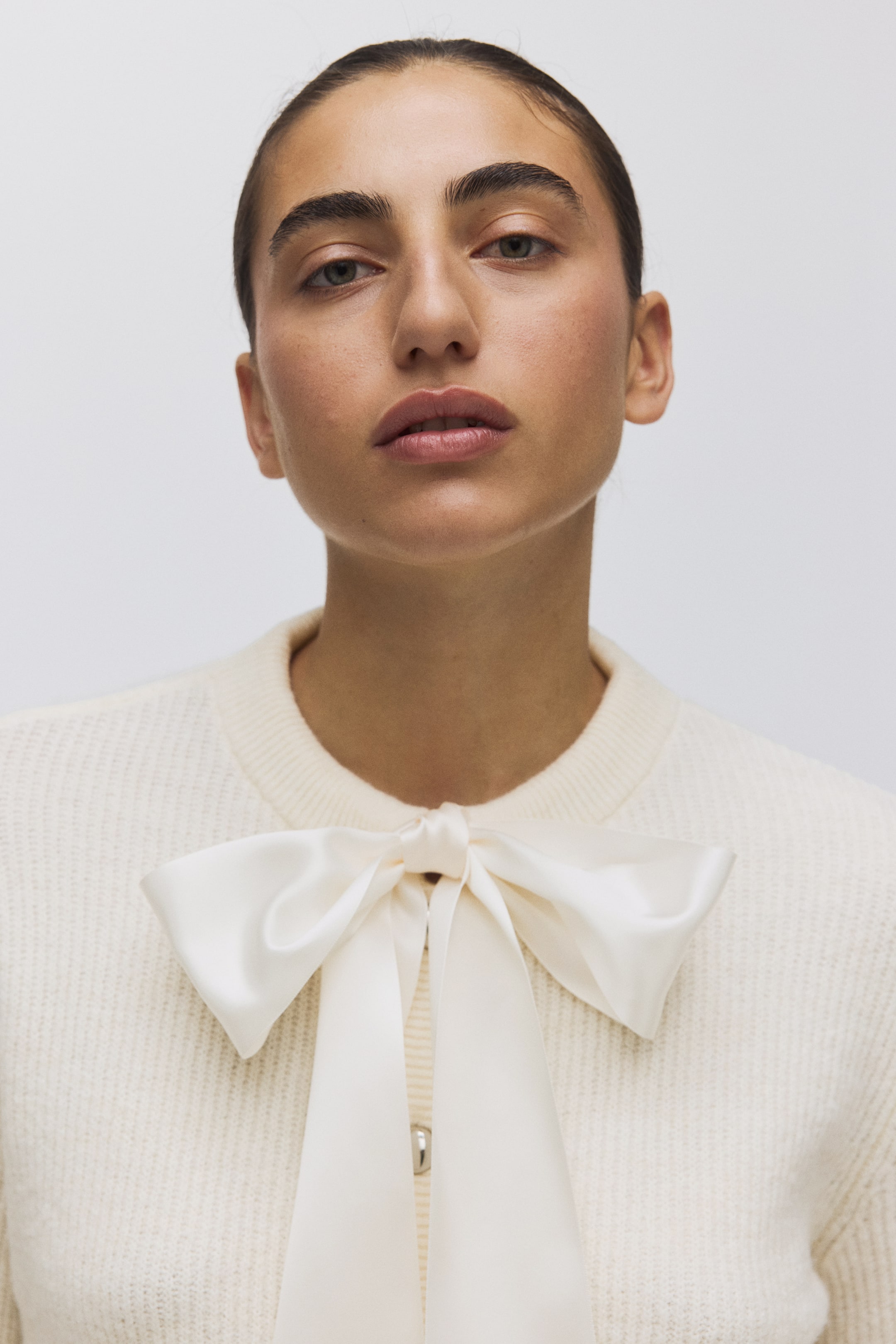 Zobacz większy obraz: Kremowy, lekki sweter zdobi okrągły dekolt oraz duża, kremowa satynowa kokarda zawiązana na środku. Srebrzysty guzik widoczny jest pod kokardą, a odzież jest zwrócona przodem.