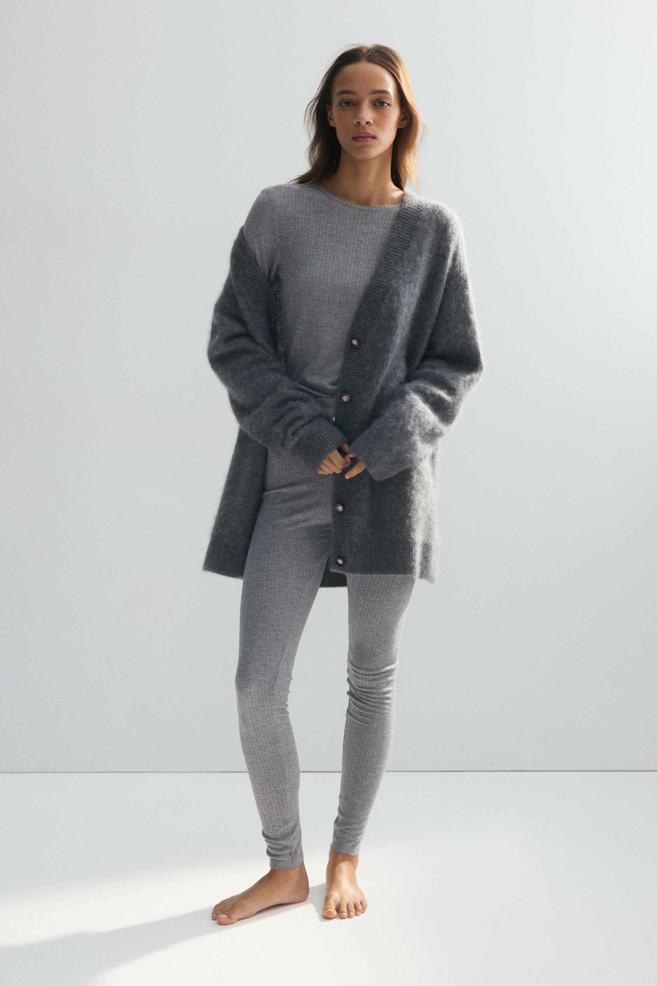 Agrandir l'image: Un ensemble de pyjama gris en jersey pointelle, composé d’un haut à manches longues et d’un legging ajusté assorti, est porté par le mannequin. L’ensemble est stylé avec un cardigan en maille gris foncé ouvert, superposé par-dessus le haut.
