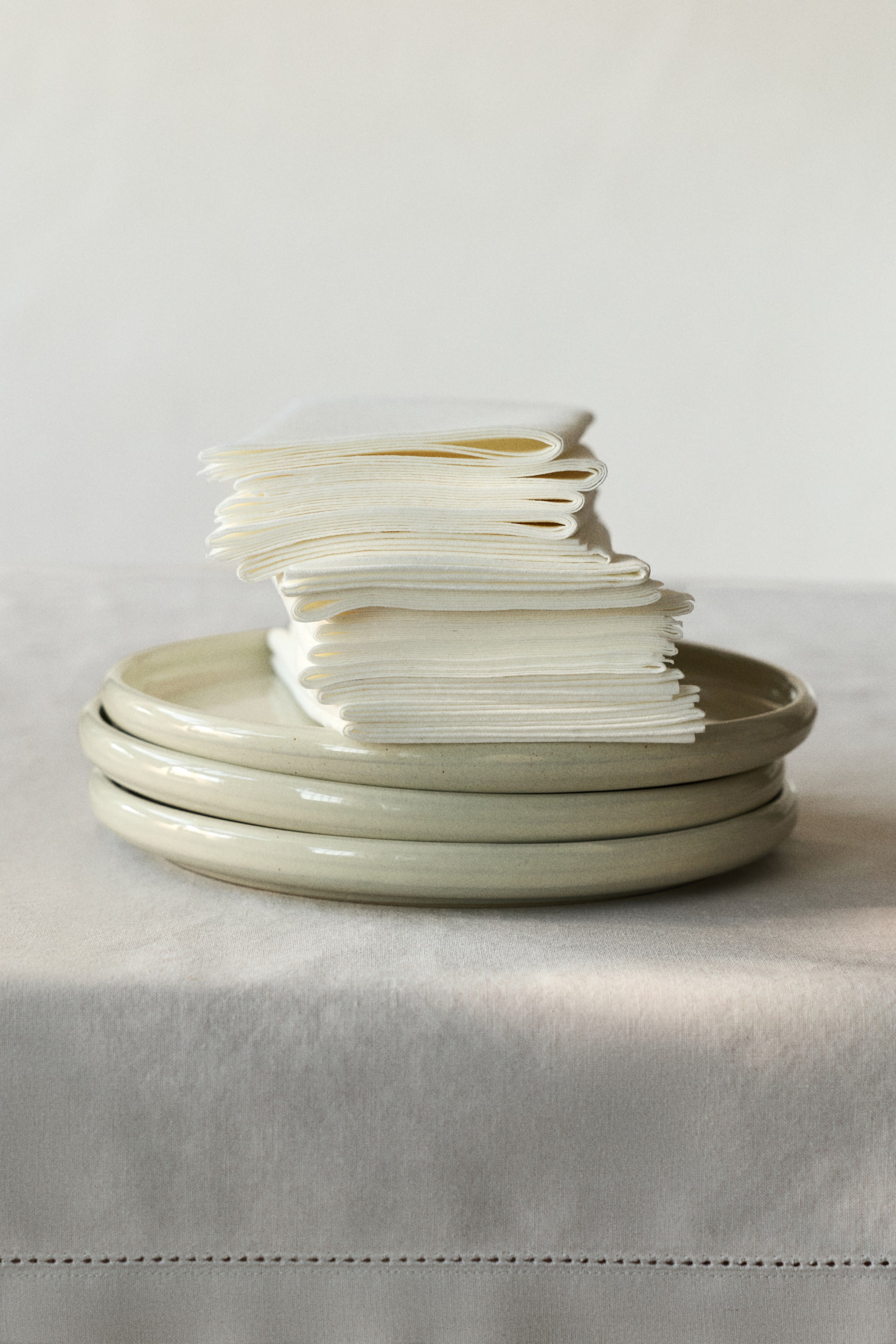 View larger image: A tall stack of white paper napkins is resting on three light beige ceramic plates. The individual napkins are neatly folded, showing their layered edges.