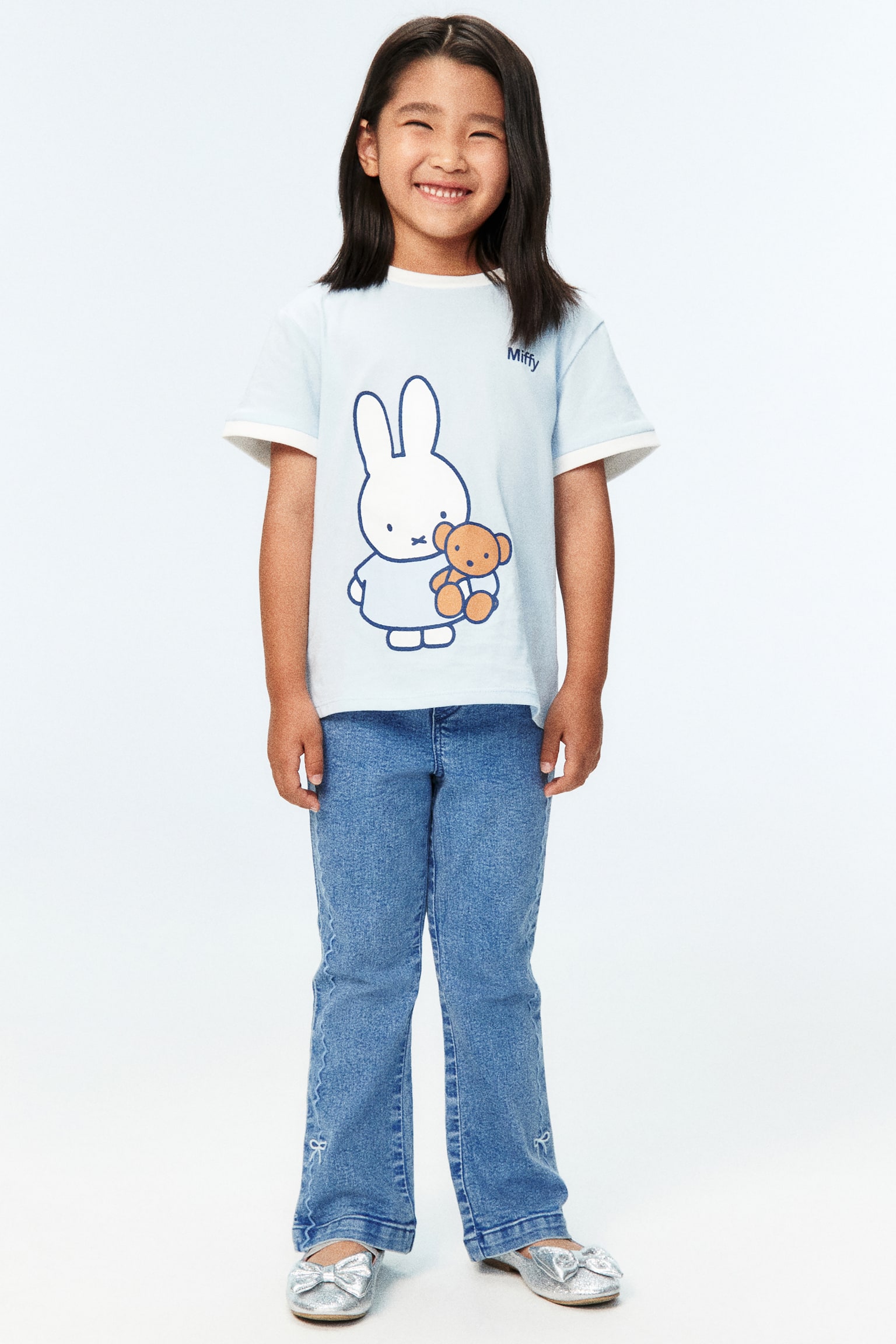 Quần jean denim xanh nhạt có cạp thun và hai túi cong phía trước. Chúng được trang trí bằng đường chỉ may xanh nhạt uốn lượn dọc theo đường may ngoài và thêu hình nơ nhỏ gần gấu quần loe. - 1