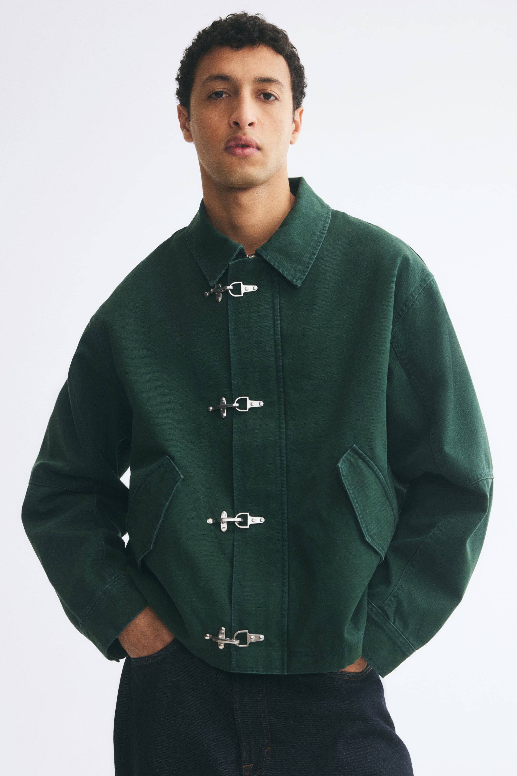 View larger image: A dark green cotton canvas jacket featuring a collar, dropped shoulders, angled flap pockets, and four silver-tone metal clasps down the front wind flap is being worn by the model. The model is standing with hands tucked into dark denim trousers.