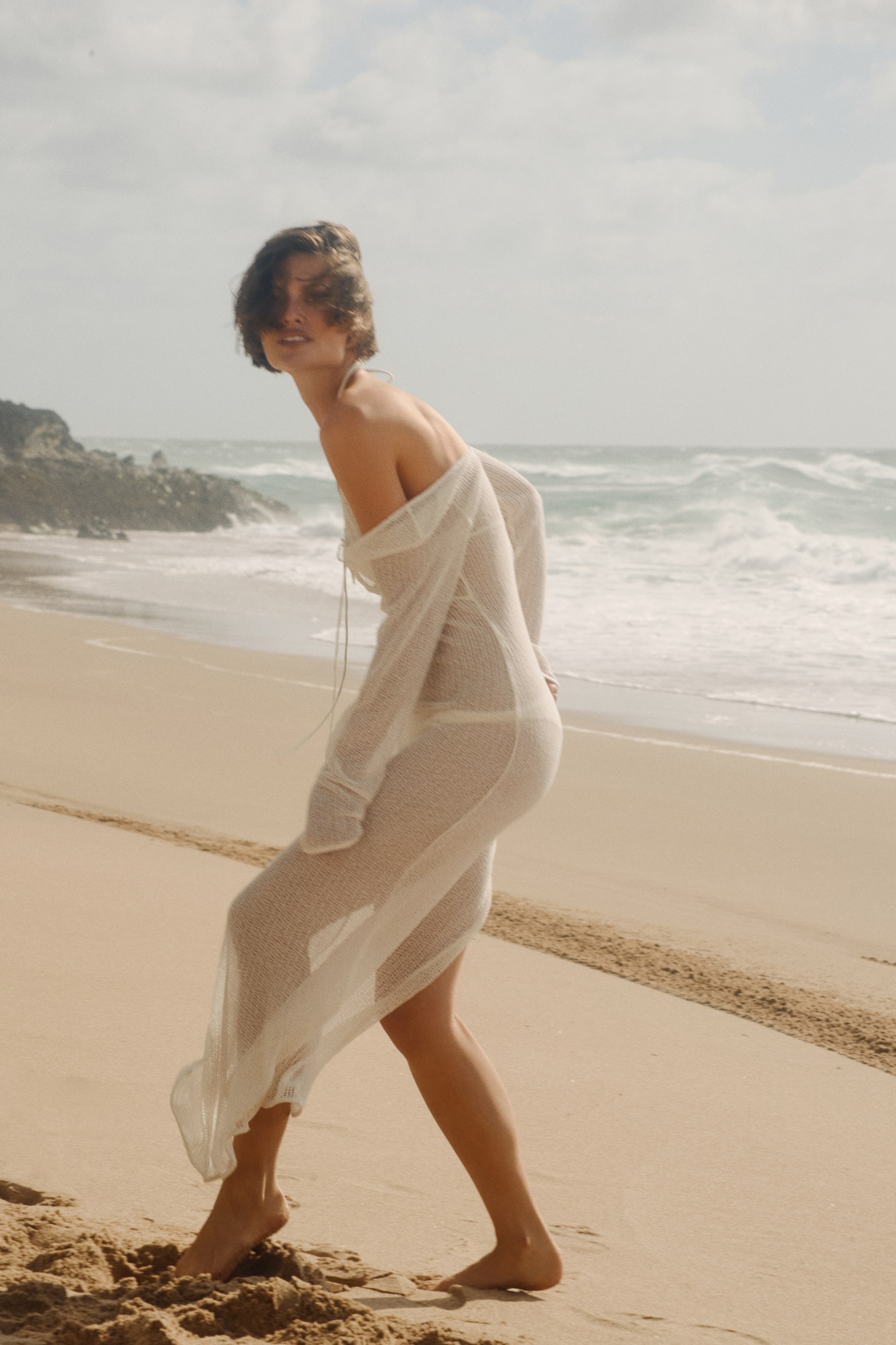 Agrandir l'image: La modèle porte une robe de plage longue, de couleur claire et à mailles lâches. Elle est dotée de manches longues, d'une texture transparente révélant un bas de bikini en dessous, et de liens étroits tombant sur la poitrine. La modèle marche, et la robe est portée de manière à découvrir les épaules, révélant une fente haute sur un côté lorsqu'elle se déplace.