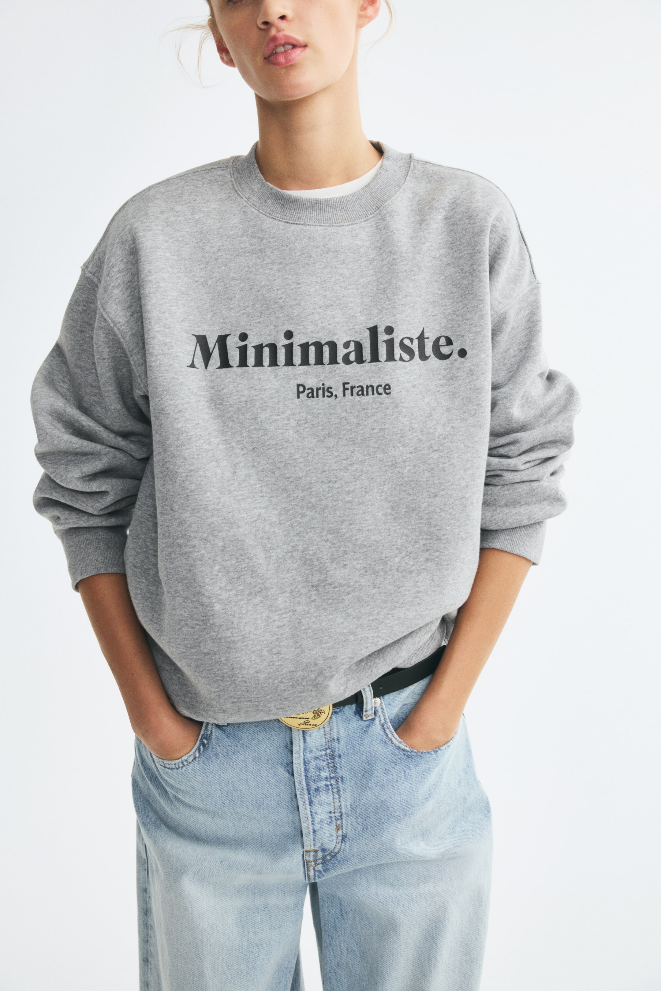 Grotere afbeelding bekijken: Een oversized grijze sweatshirt met een geribbelde ronde hals, verlaagde schouders en de zwarte tekst 'Minimaliste. Paris, France' op de borst wordt door het model gedragen. Het model staat met de handen in de zakken van een lichtgewassen denim jeans.