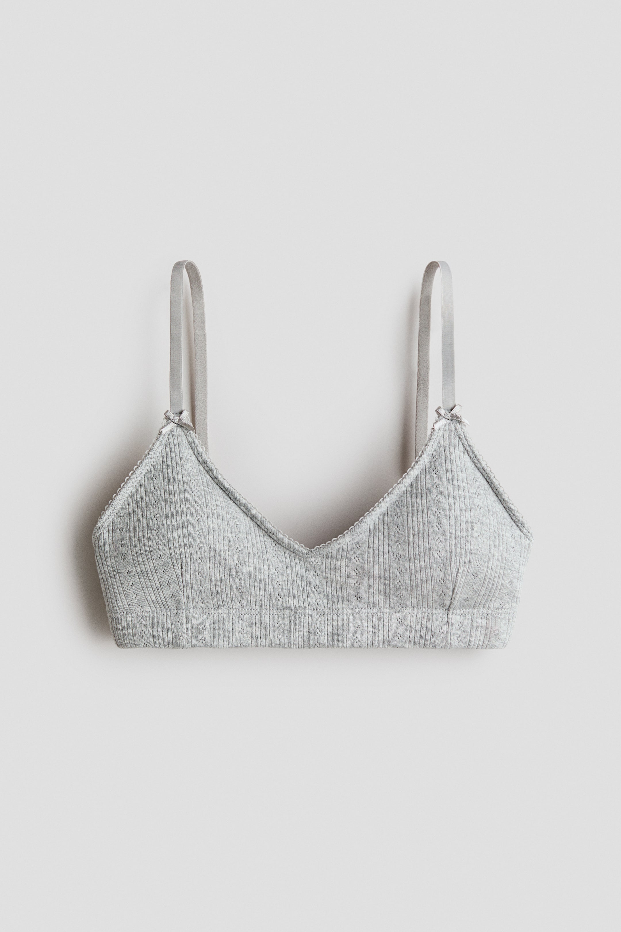 Ver imagen más grande: Un top corto gris se presenta desde el frente, con un escote en forma de V adornado con un ribete de picot. Está diseñado con tirantes estrechos y ajustables que están decorados con pequeños lazos de satén, y muestra una textura suave de jersey de punto de algodón acanalado.