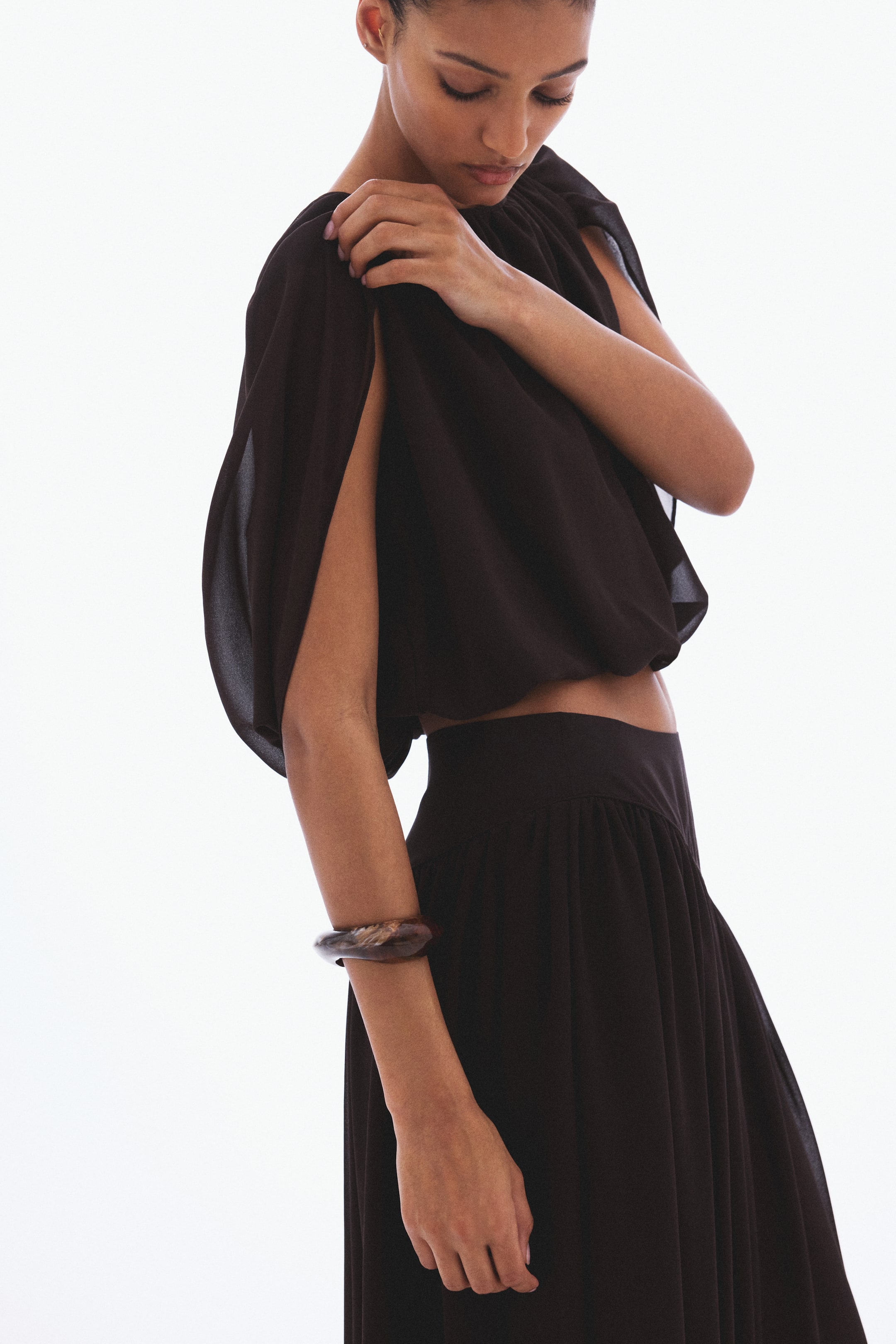 View larger image: A black cropped top features a wide, draped neckline with flowing, open sleeves that reveal the arm through its side openings. A hand holds the top's shoulder, emphasizing the fabric's drape. It's paired with a matching high-waisted, flowing skirt, gathered at the waist.