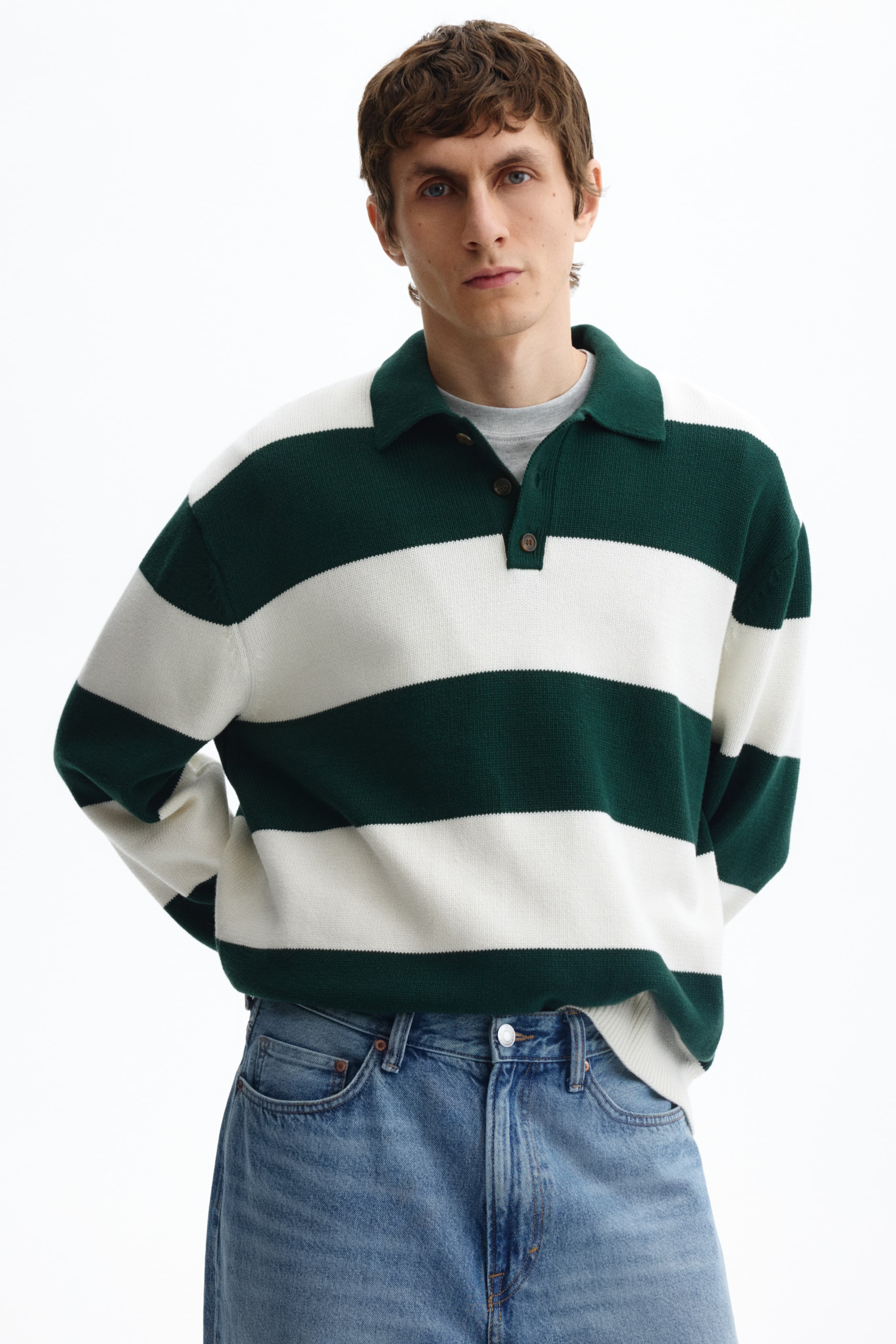 Ver imagen más grande: Un polo de punto con anchas rayas horizontales en verde oscuro y blanco, diseñado con un cuello de pico en verde oscuro y una tapeta con dos botones marrones. Presenta mangas largas y puños acanalados, y se aprecia una camiseta de color gris claro bajo la tapeta.
