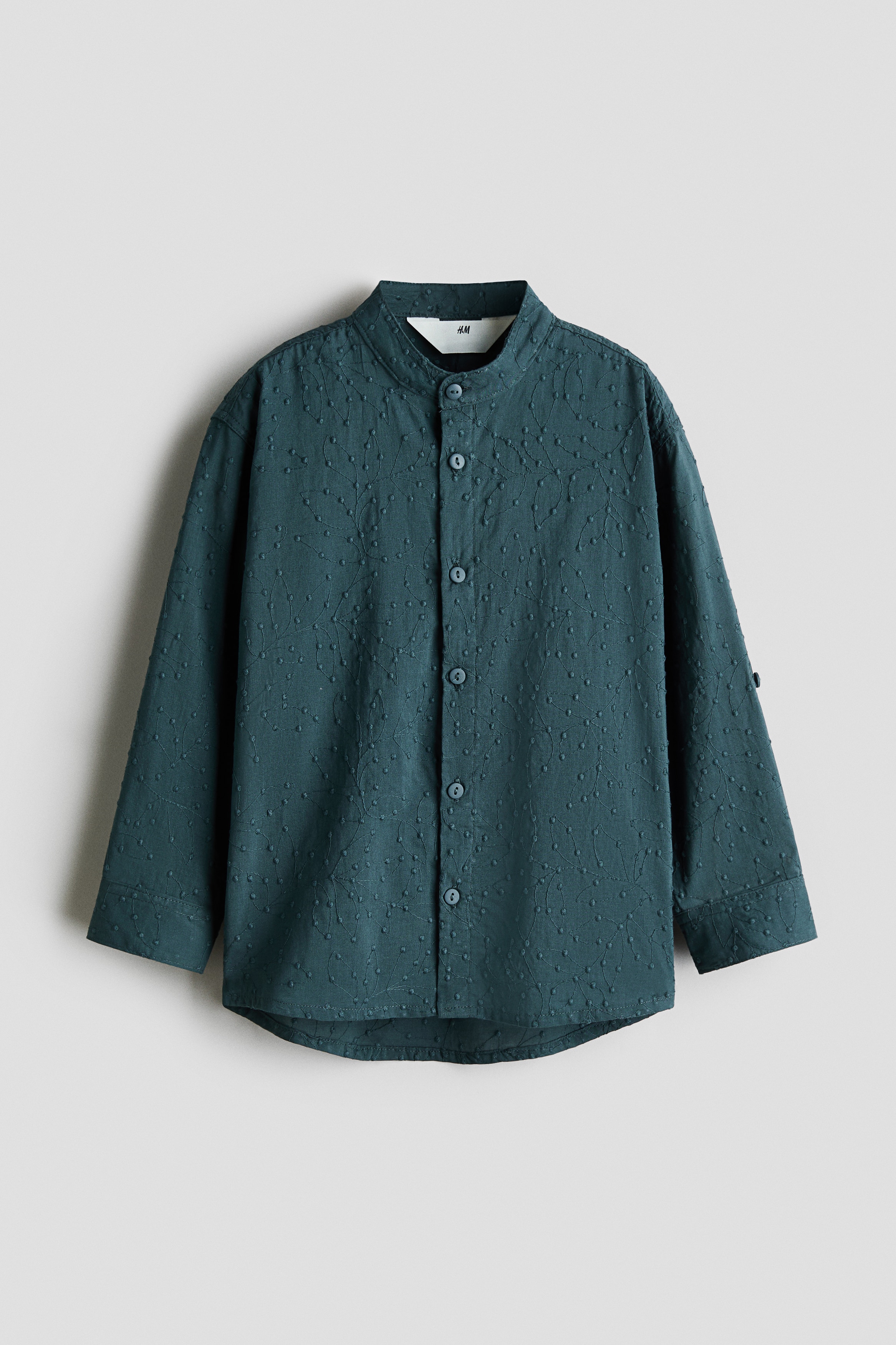 A dark teal grandad shirt in a patterned cotton weave is shown from the front, featuring a band collar and matching buttons down the front. The long sleeves are rolled up on the arms, displaying the button cuffs and button tabs.
