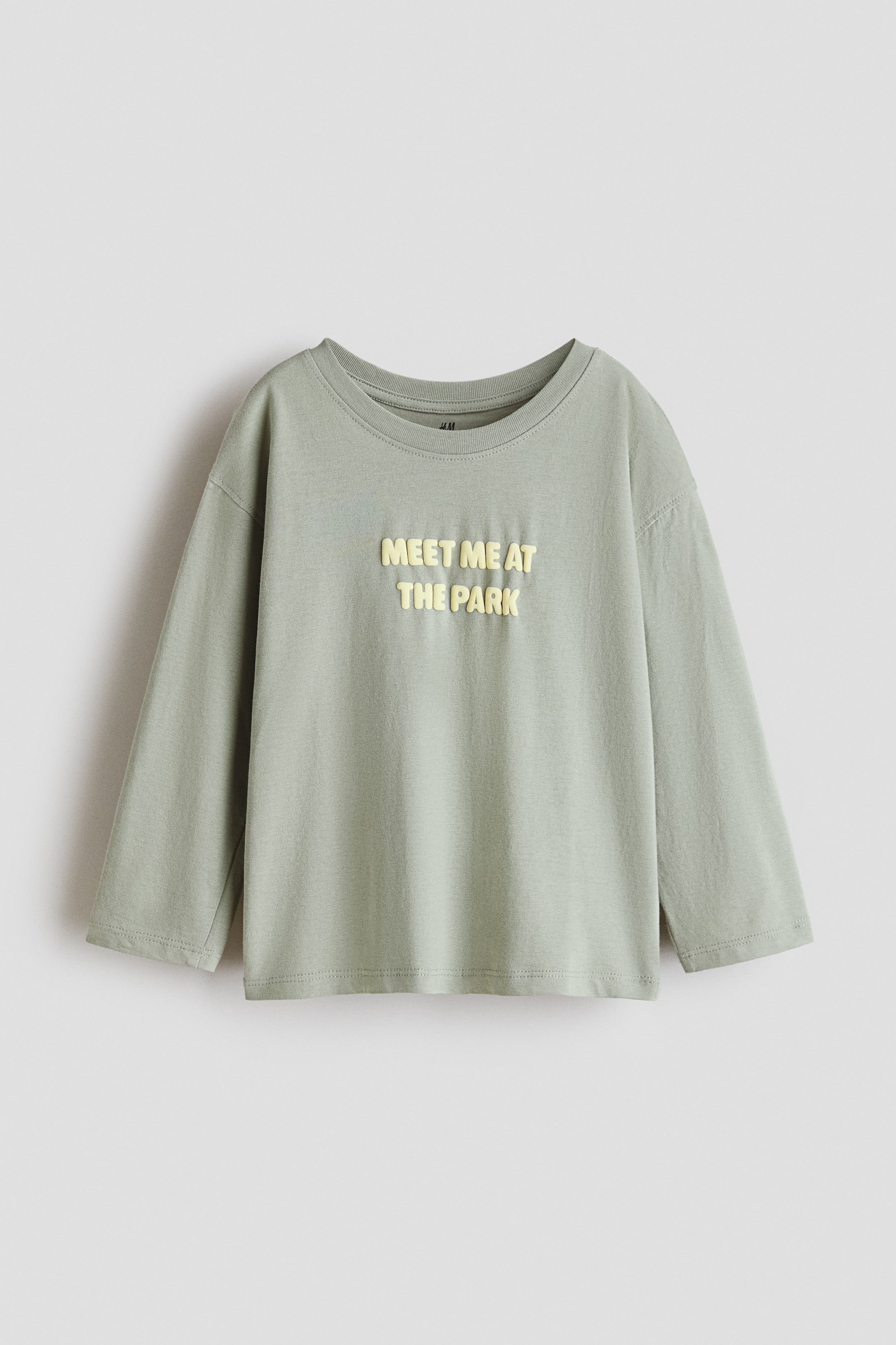 Voir une image plus grande: Un T-shirt à manches longues vert pâle en jersey de coton est posé à plat, présentant un col rond bordé et des épaules tombantes. Une inscription en relief jaune pâle sur la poitrine affiche « MEET ME AT THE PARK ». 