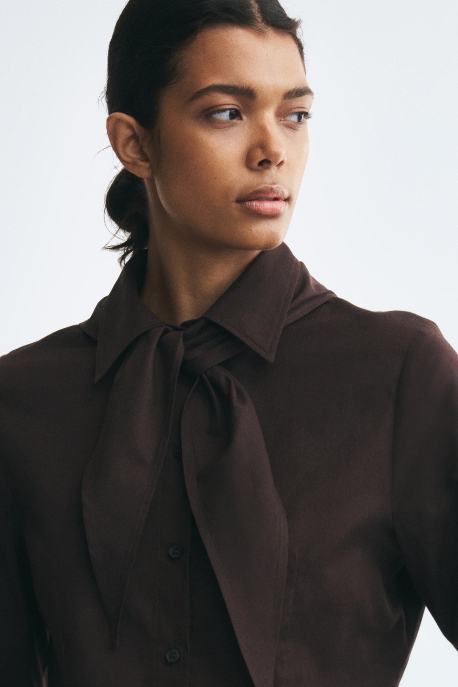 The model is wearing a dark brown cotton shirt, featuring a pointed collar with a matching scarf tied around the neck and a button-up front. - 1