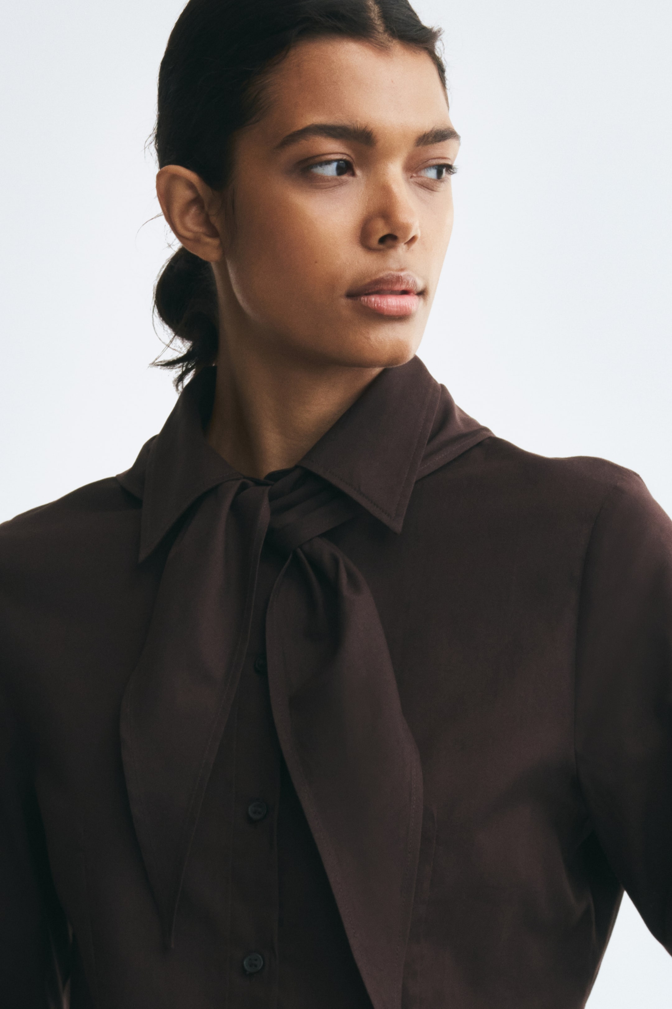 View larger image: The model is wearing a dark brown cotton shirt featuring a pointed collar, a matching scarf tied at the neck, and a button-front closure.