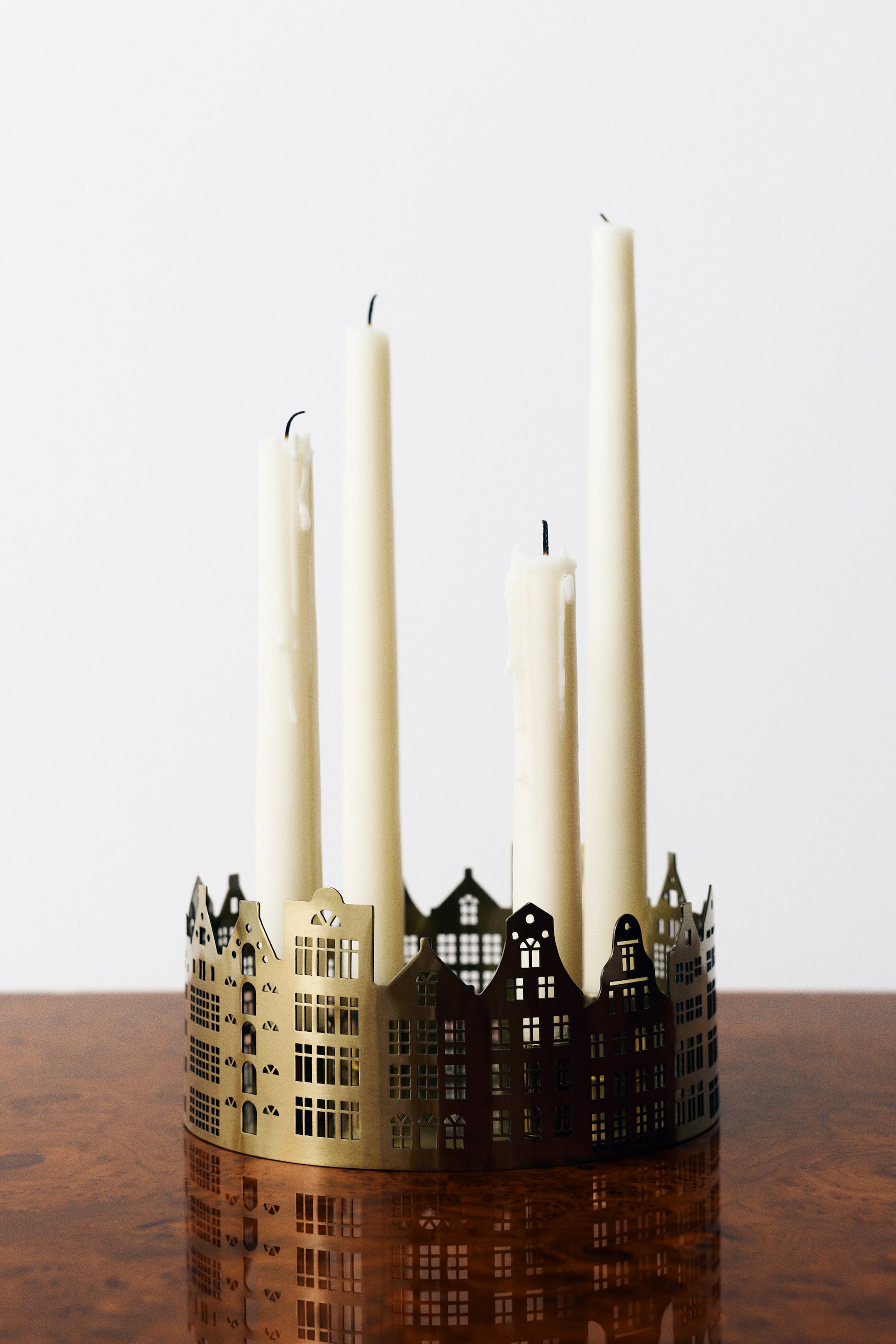 View larger image: Shown from the front, a round, gold-toned metal candleholder featuring a cut-out cityscape design with detailed windows and gabled roofs holds four cream-coloured taper candles of varying heights.