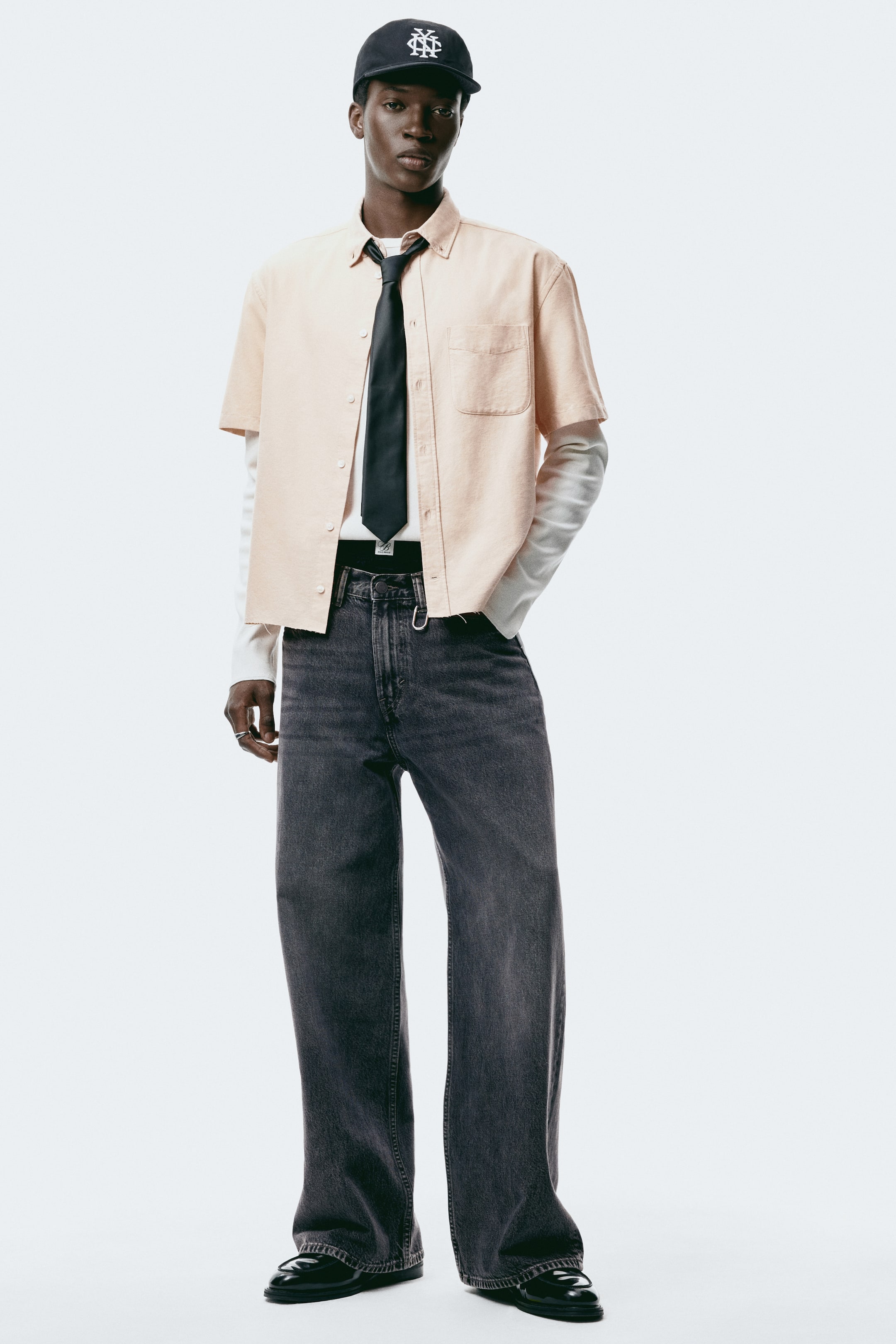 Ver imagem maior: Uma camisa de botões de manga curta em pêssego claro apresenta uma gola abotoável, um bolso aplicado no peito esquerdo e uma bainha sem rematar. Está usada com a gola desabotoada, com uma gravata preta, e sobreposta a uma camisa branca de manga comprida.