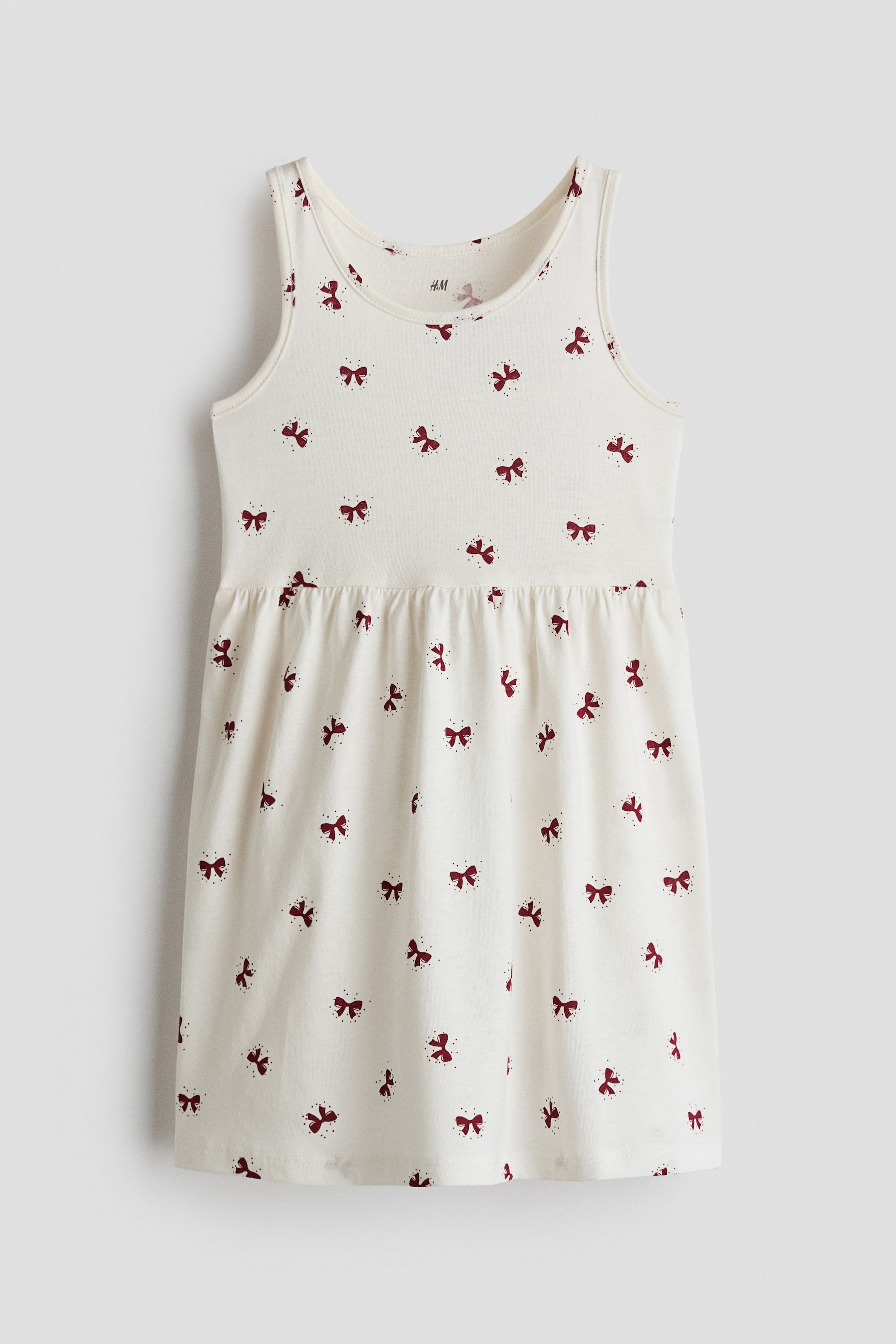 View larger image: A sleeveless, off-white cotton jersey dress is shown from the front, featuring an all-over print of small burgundy bows. The dress features a round neckline, a gathered seam at the waist, and a flared skirt.