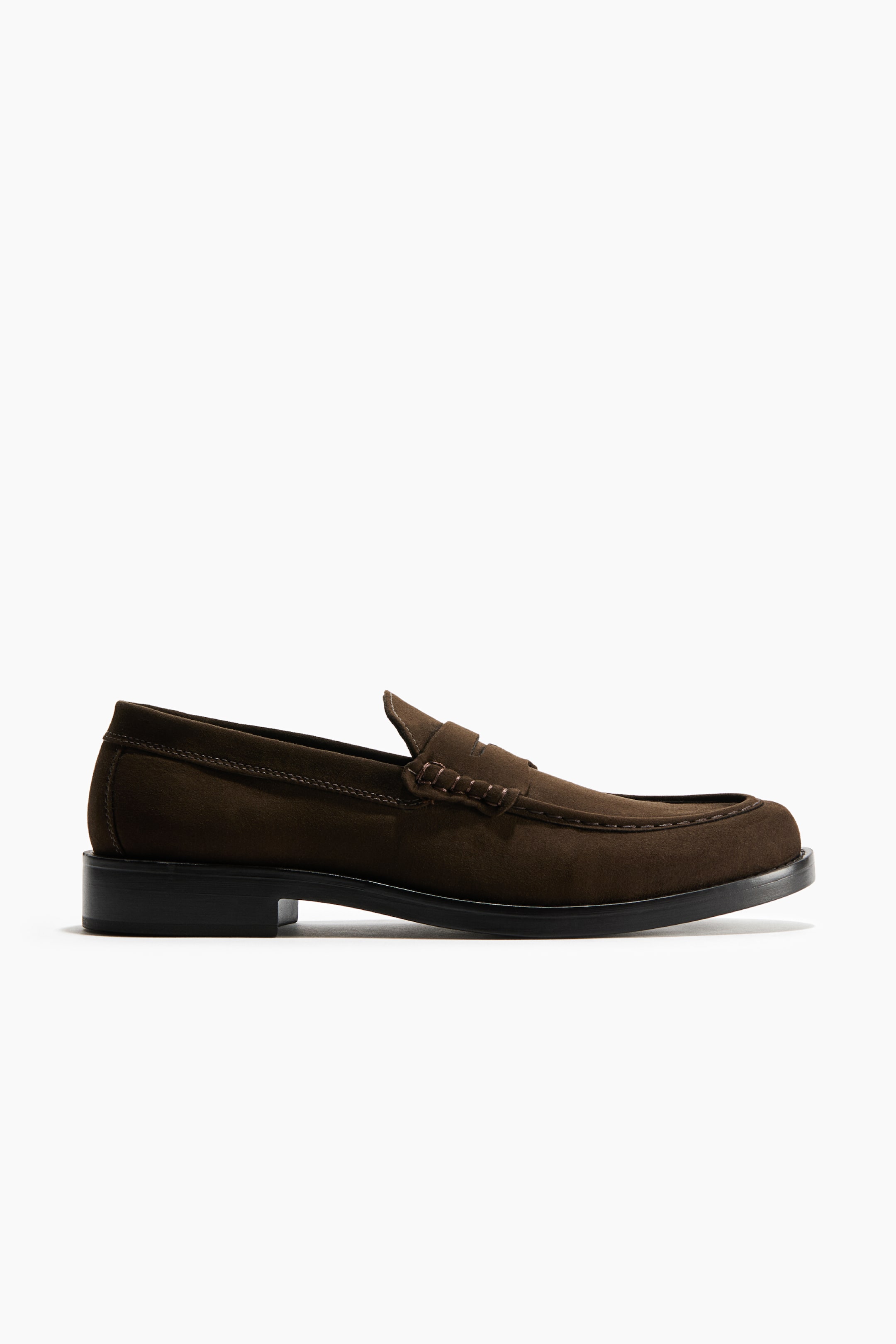 View larger image: A dark brown suede penny loafer is shown in a side profile view, featuring a classic saddle strap with beefroll stitching across the vamp and a stitched apron toe. The shoe rests on a low block heel and a thick black sole.
