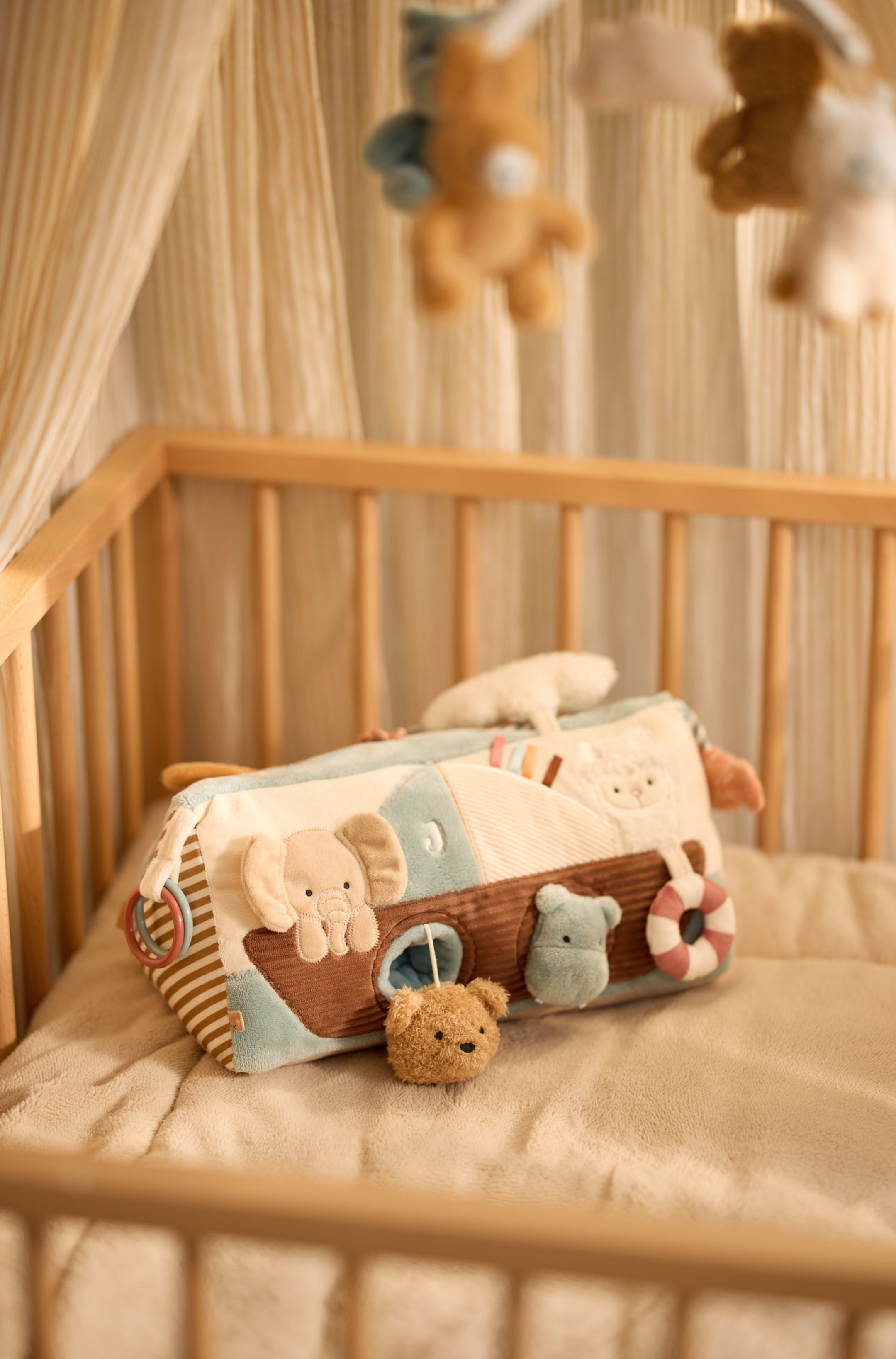 Agrandir l'image: Un jouet d’éveil en peluche doux, de forme bateau, est posé en biais, avec différents panneaux aux couleurs pastel, du velours côtelé texturé et des figurines d’animaux intégrées, dont un éléphant et un lama. Une petite tête d’ours en peluche est suspendue par un cordon à partir d’une ouverture sur le côté, tandis que des anneaux en plastique pendent à une extrémité.