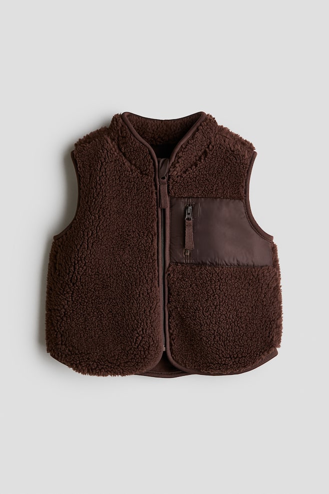 Ein brauner, weicher Teddy-Gilet wird von vorne gezeigt, mit einem Stehkragen, einem durchgehenden Frontreißverschluss mit einem scheuerfreien Kinnschutz und einer Reißverschlusstasche aus gewebtem Stoff auf der linken Brust, alles mit Jersey-Einfassungen versehen. - 1