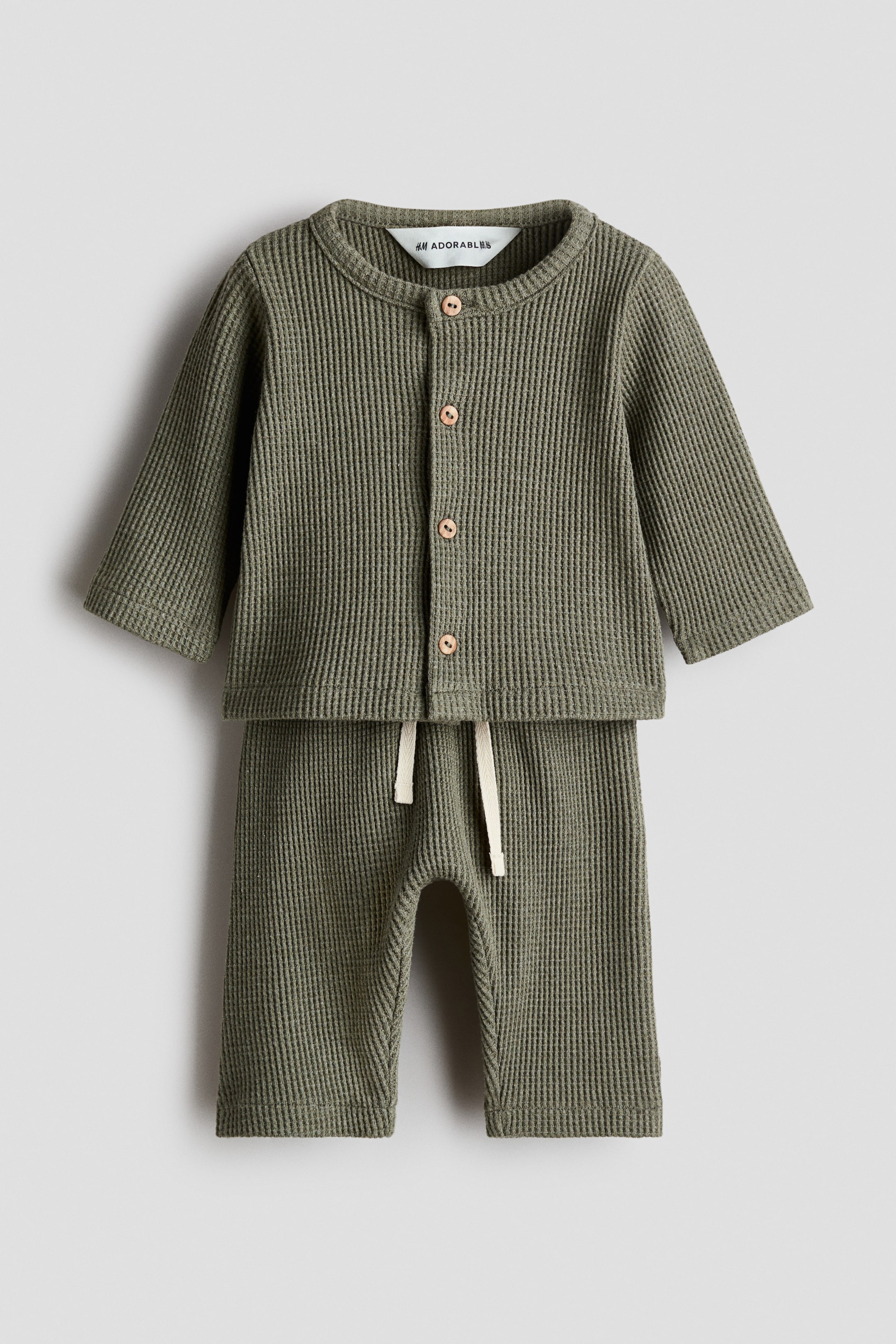 View larger image: A baby's two-piece set in green waffled jersey is displayed, featuring a cardigan with a round neckline and wooden buttons down the front, and trousers with a cream drawstring at the waist.
