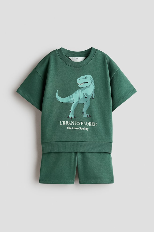 A front view of a green two-piece loungewear set featuring a short-sleeved sweatshirt and matching shorts. The sweatshirt is displaying a light-blue dinosaur graphic with the text "URBAN EXPLORER" and "The Dino Society" printed below. - 1