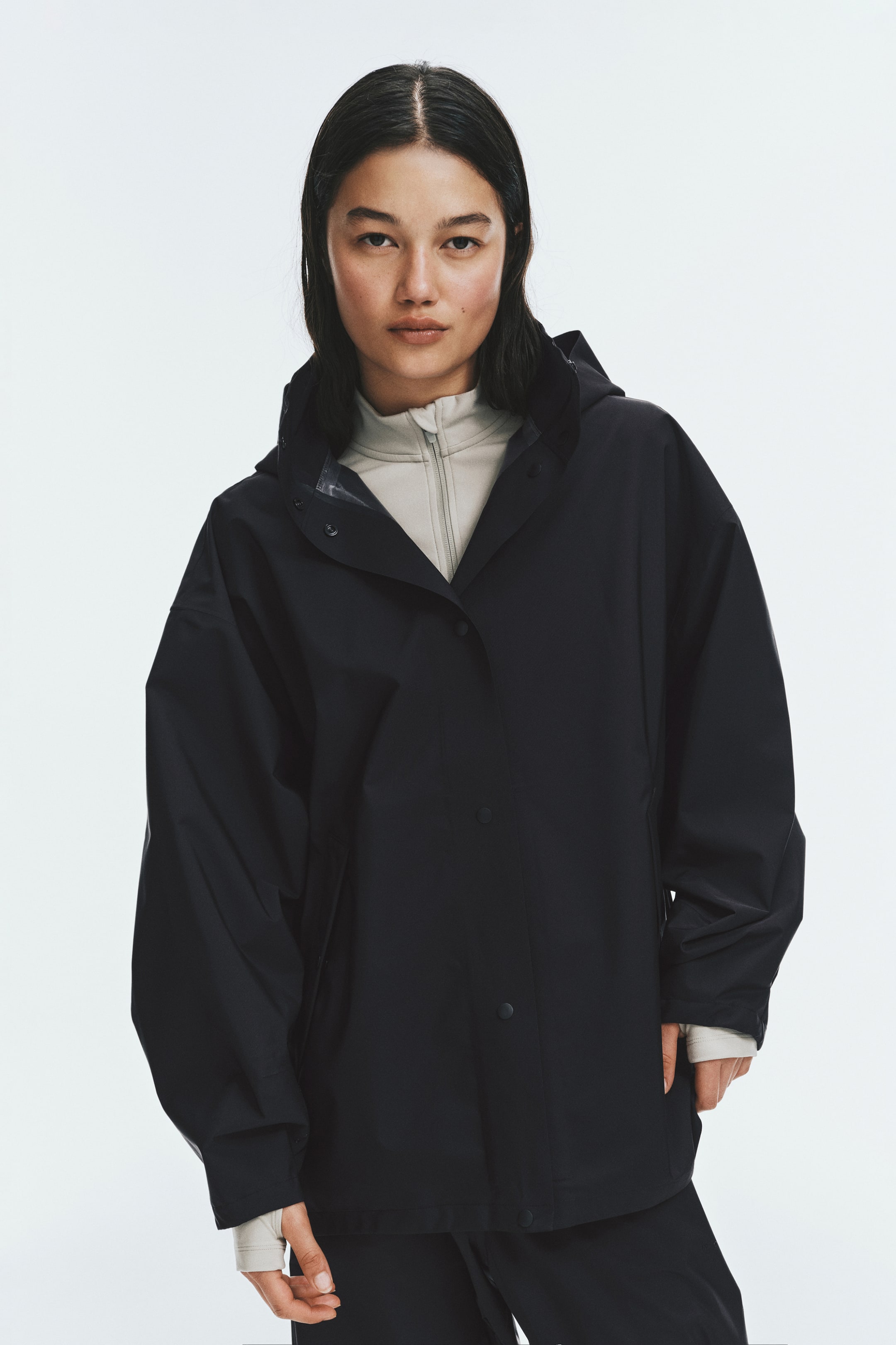 Visualizza immagine più grande: Una giacca nera con cappuccio, dalla vestibilità oversize e con chiusure a pressione, indossata dalla modella. La giacca è sovrapposta a un top con collo alto e zip di colore chiaro ed è abbinata a pantaloni neri coordinati.