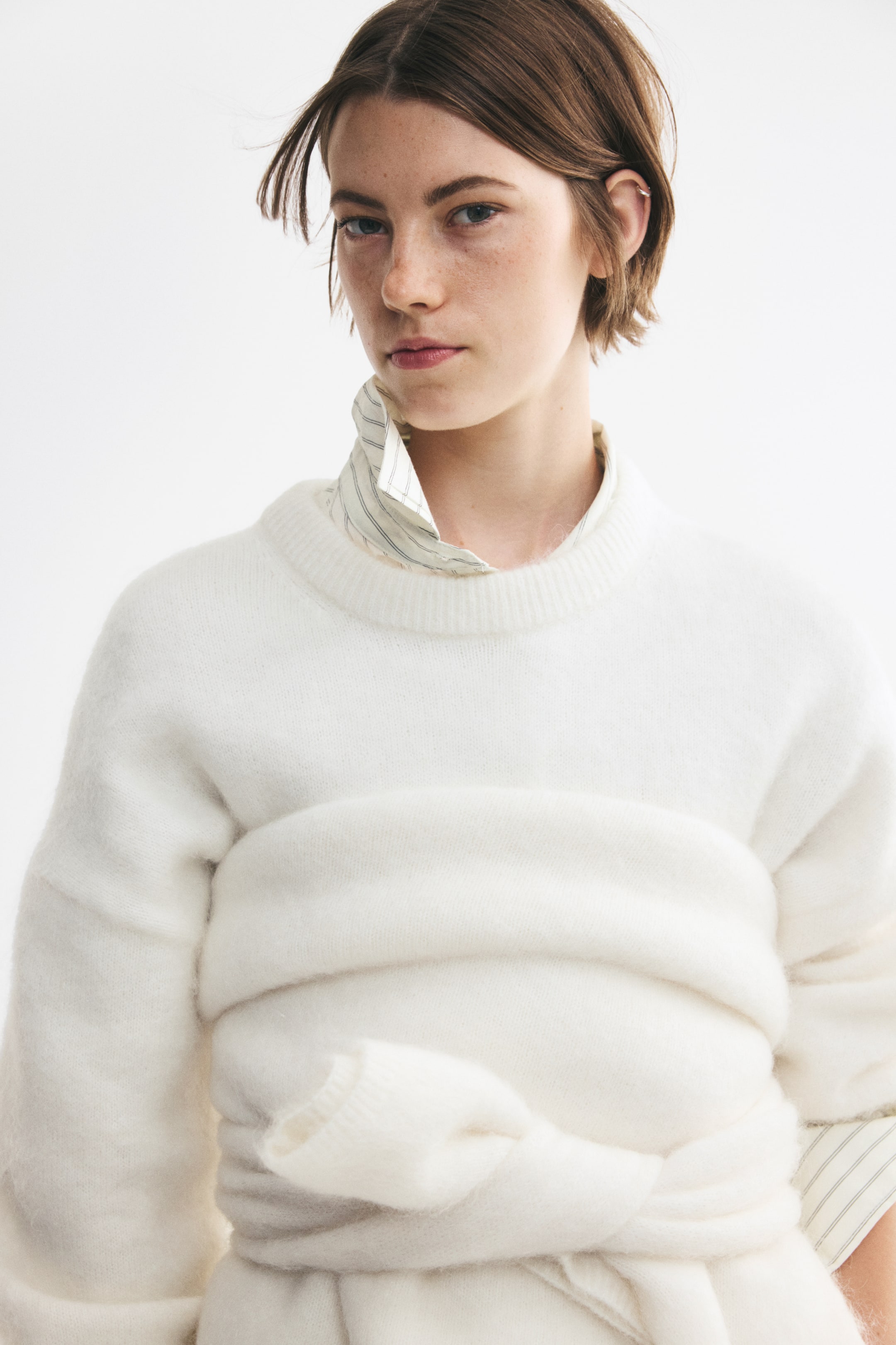 Grotere afbeelding bekijken: Een oversized crèmekleurige gebreide trui presenteert een zachte, pluizige textuur met een geribde ronde hals en verlaagde schouders in een vooraanzicht. De trui wordt gestyled in laagjes over een gestreept overhemd met kraag, met een tweede bijpassende trui om de taille gewikkeld en vastgeknoopt.