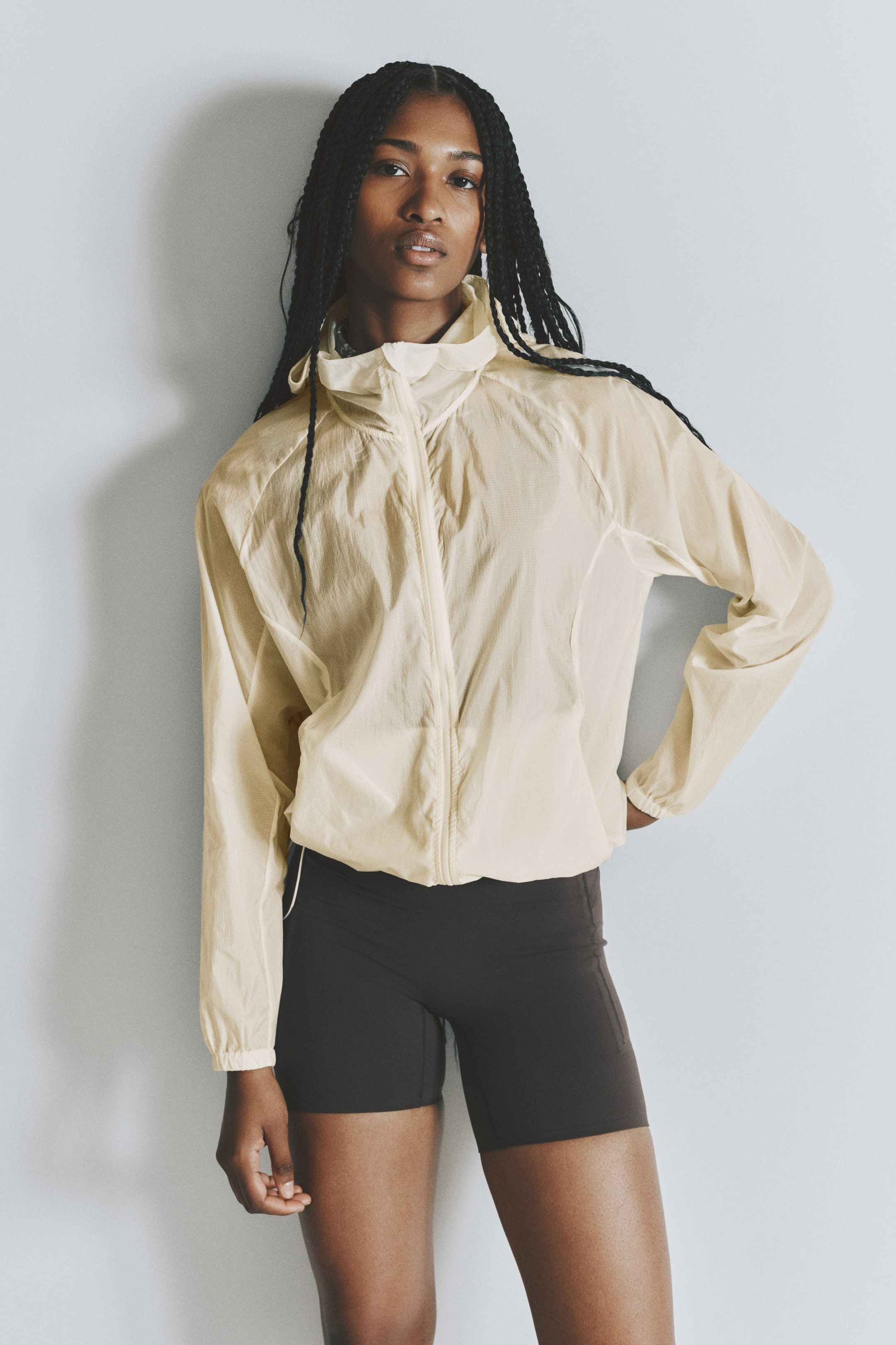 View larger image: A pale yellow translucent windbreaker jacket, featuring a high collar, full front zipper, and elasticated cuffs and hem, is being worn over dark brown biker shorts.