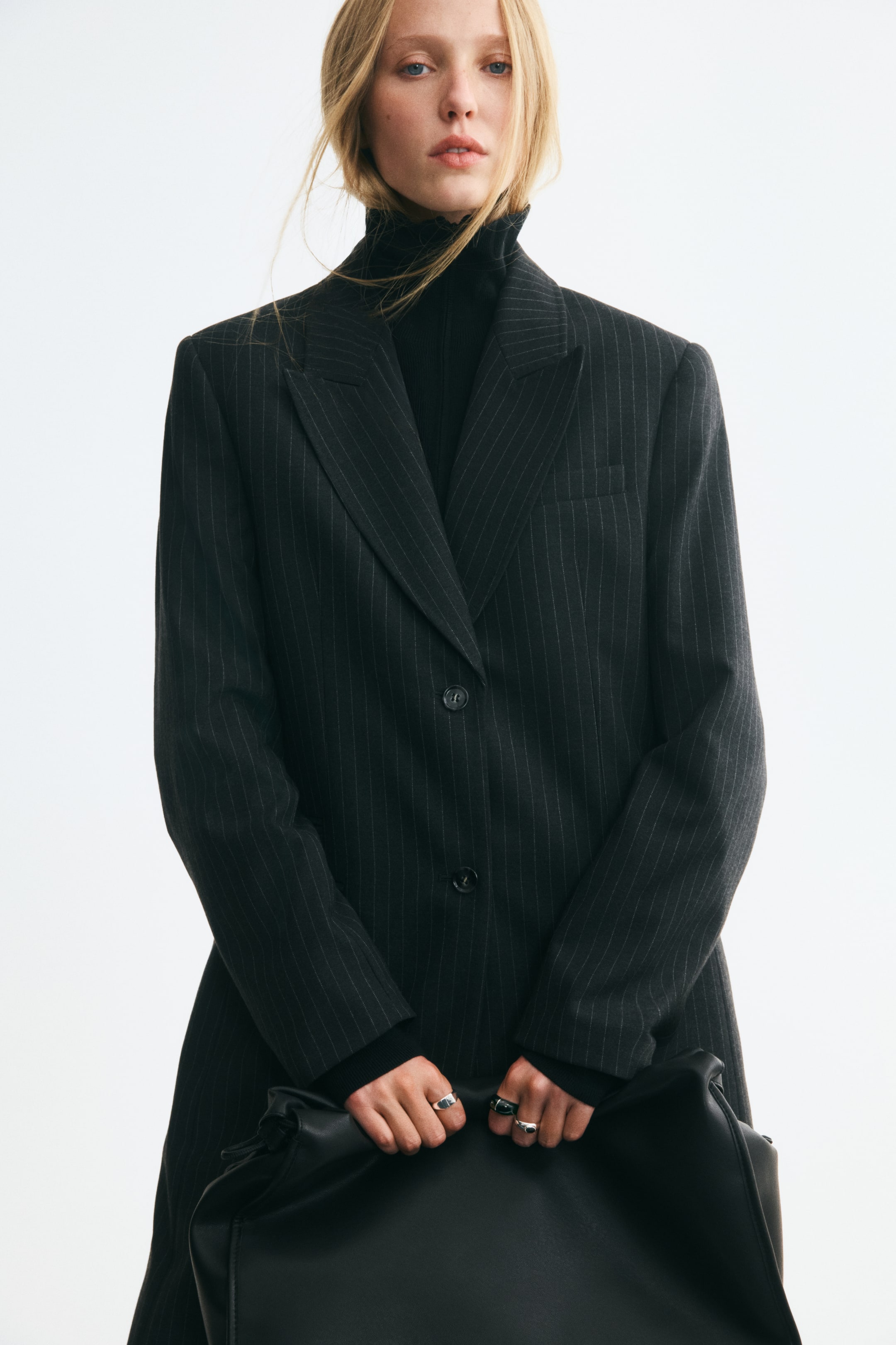 Ver imagem maior: A modelo veste um blazer de trespasse simples em cinzento escuro às riscas finas sobre uma camisola preta de gola alta. O blazer oversized apresenta lapelas em bico, fecho frontal com botões e ombros estruturados, e é complementado por uma mala grande preta segurada à frente.