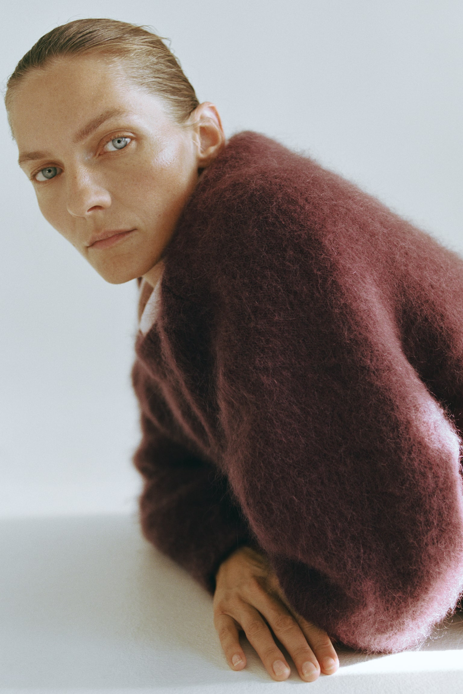 A fuzzy burgundy mohair-blend sweater is worn by the model, who is leaning forward onto a flat surface. The oversized sweater features a soft, textured knit, dropped shoulders and a slightly visible white collar layered underneath. - 1