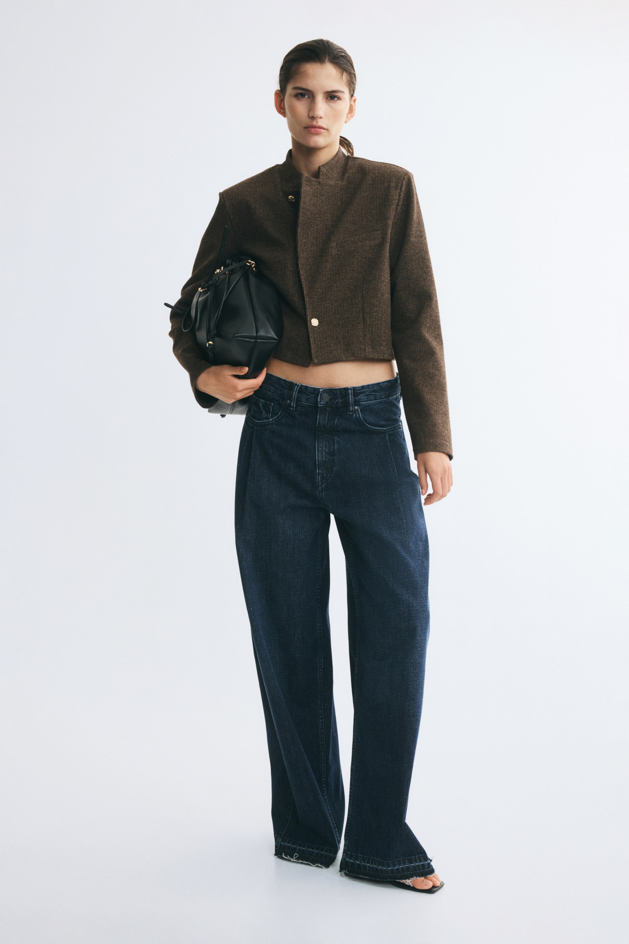 이미지 확대 보기: 텍스처가 있는 직조 원단으로 제작된 크롭 기장의 루즈핏 브라운 블레이저를 모델이 착용하고 있으며, 스탠드 칼라, 구조적인 어깨 라인, 그리고 두 개의 버튼이 보이는 오프셋 프런트 클로저가 특징입니다. 블레이저는 짙은 블루 컬러의 와이드 레그 진과 매치되었고, 블랙 핸드백은 모델의 팔 아래에 들려 있습니다.