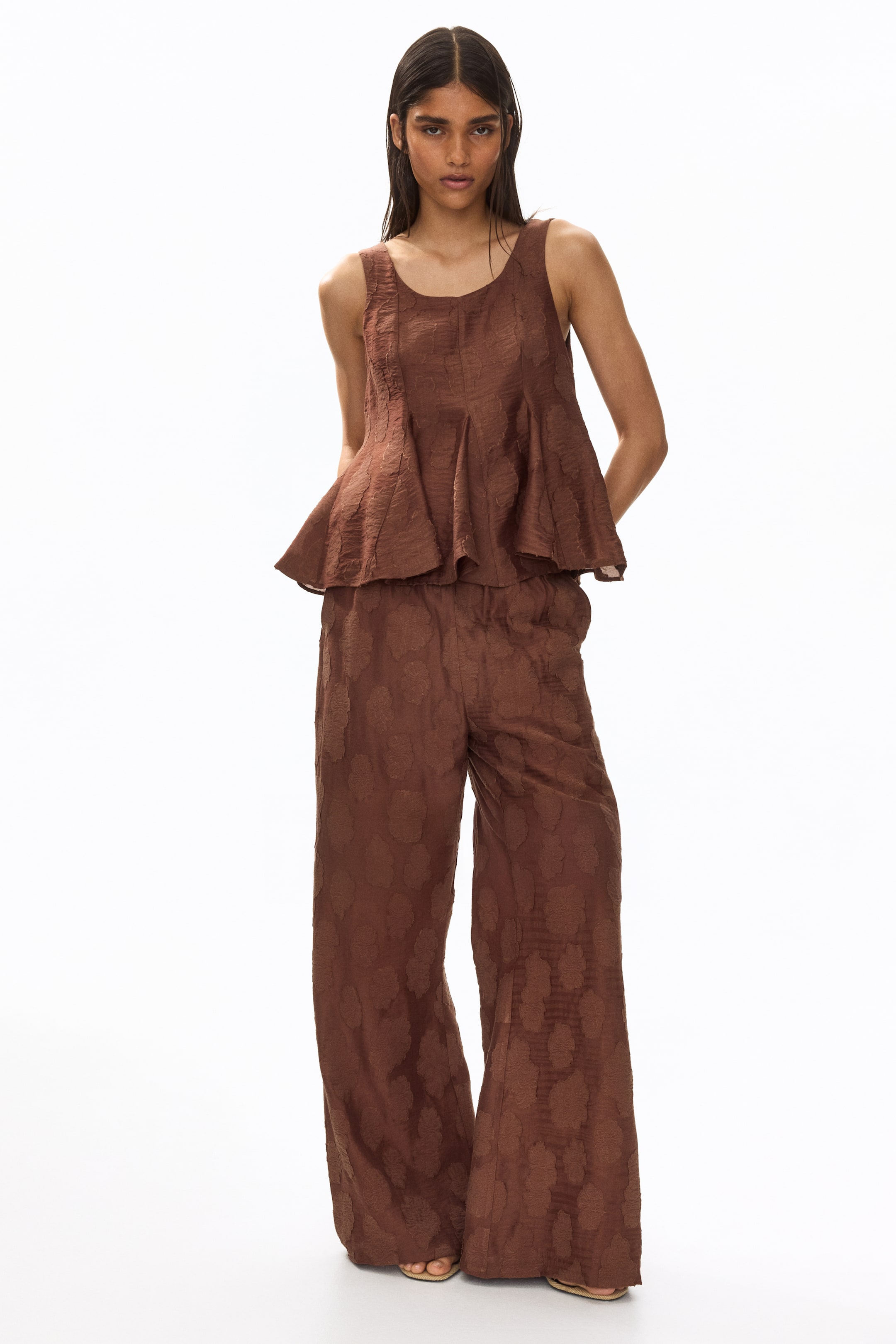 View larger image: The model is wearing brown fitted trousers in a jacquard weave, featuring an elasticated waist, diagonal side pockets, and flared legs.