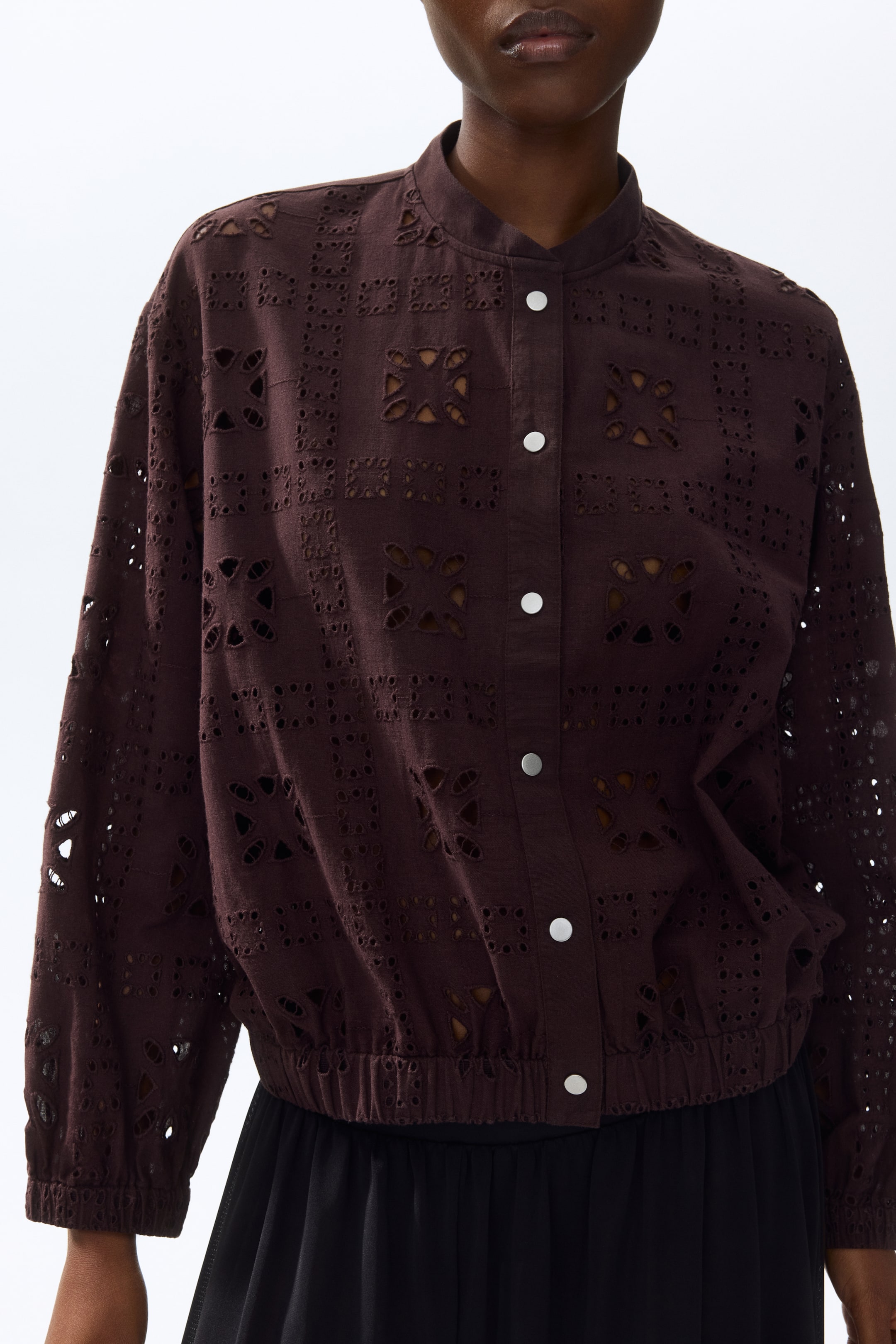 Ver imagen más grande: Se presenta una chaqueta marrón oscuro desde el frente, con un patrón de ojales geométricos en toda la superficie, un cuello tipo banda y una tapeta frontal con botones a presión de tono plateado. Exhibe mangas largas con puños elásticos y un dobladillo elástico, creando una silueta blusón.