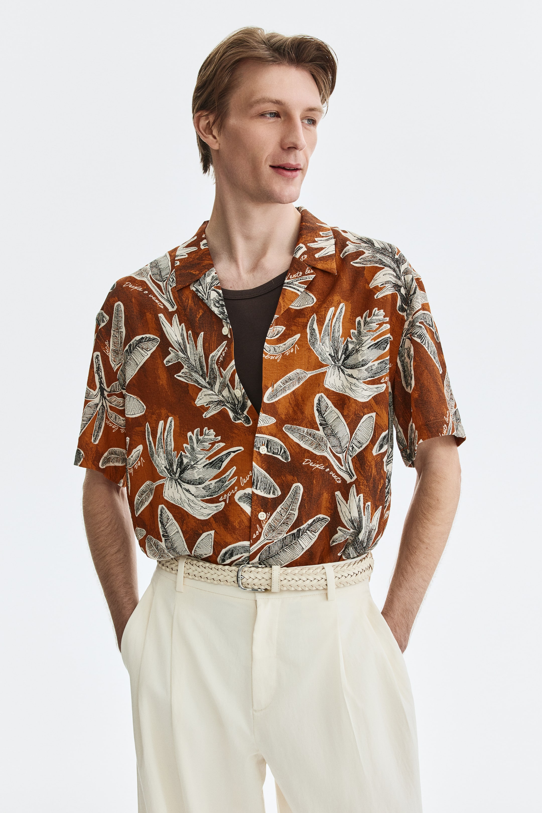 View larger image: The model is donning a short-sleeved shirt crafted from a breathable, brown and white foliage-patterned weave. This piece features a resort collar, a French front, and a straight-cut hem. Designed with a relaxed fit, the shirt is styled open over a dark undershirt, offering a casual yet chic look.