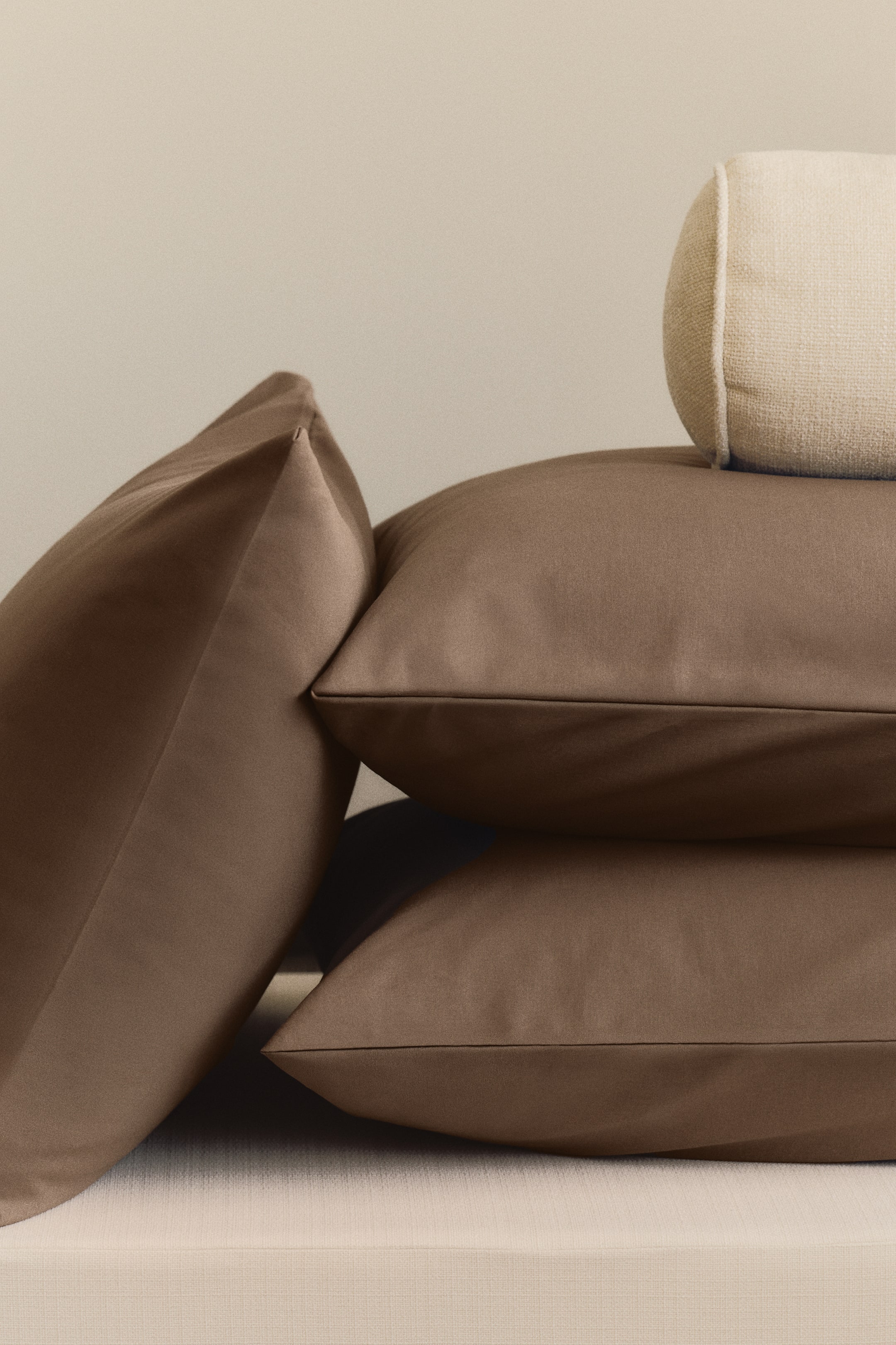 Agrandir l'image: Trois coussins rectangulaires marron sont disposés, deux empilés horizontalement et un s'appuyant contre eux depuis la gauche. Un coussin cylindrique beige clair avec un tissu texturé repose sur le coussin marron du dessus, incliné depuis la droite.