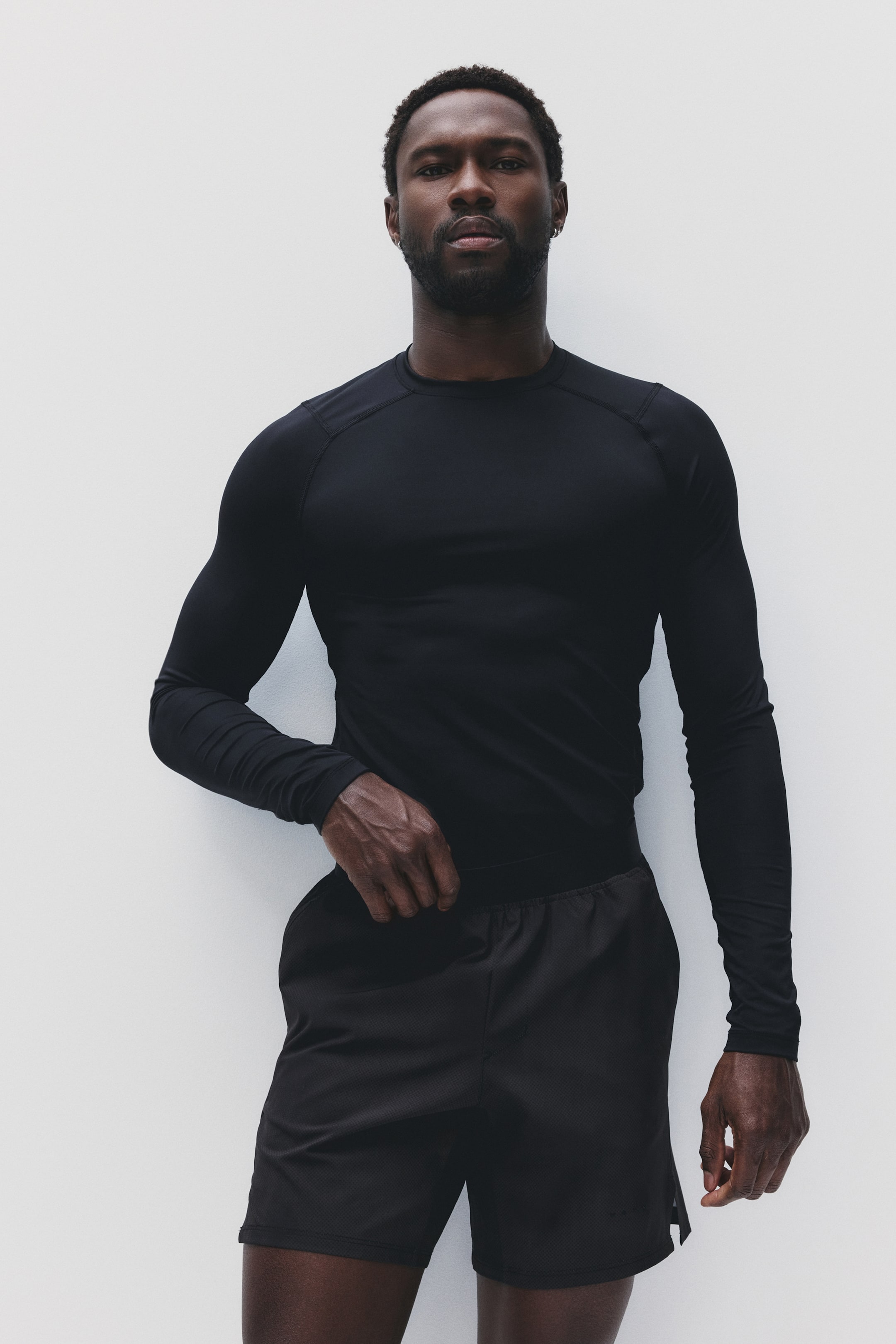 View larger image: A black, form-fitting long-sleeve athletic shirt with a crew neck and raglan sleeves showcasing visible stitching is paired with black athletic shorts that feature a subtle textured pattern and a side split.