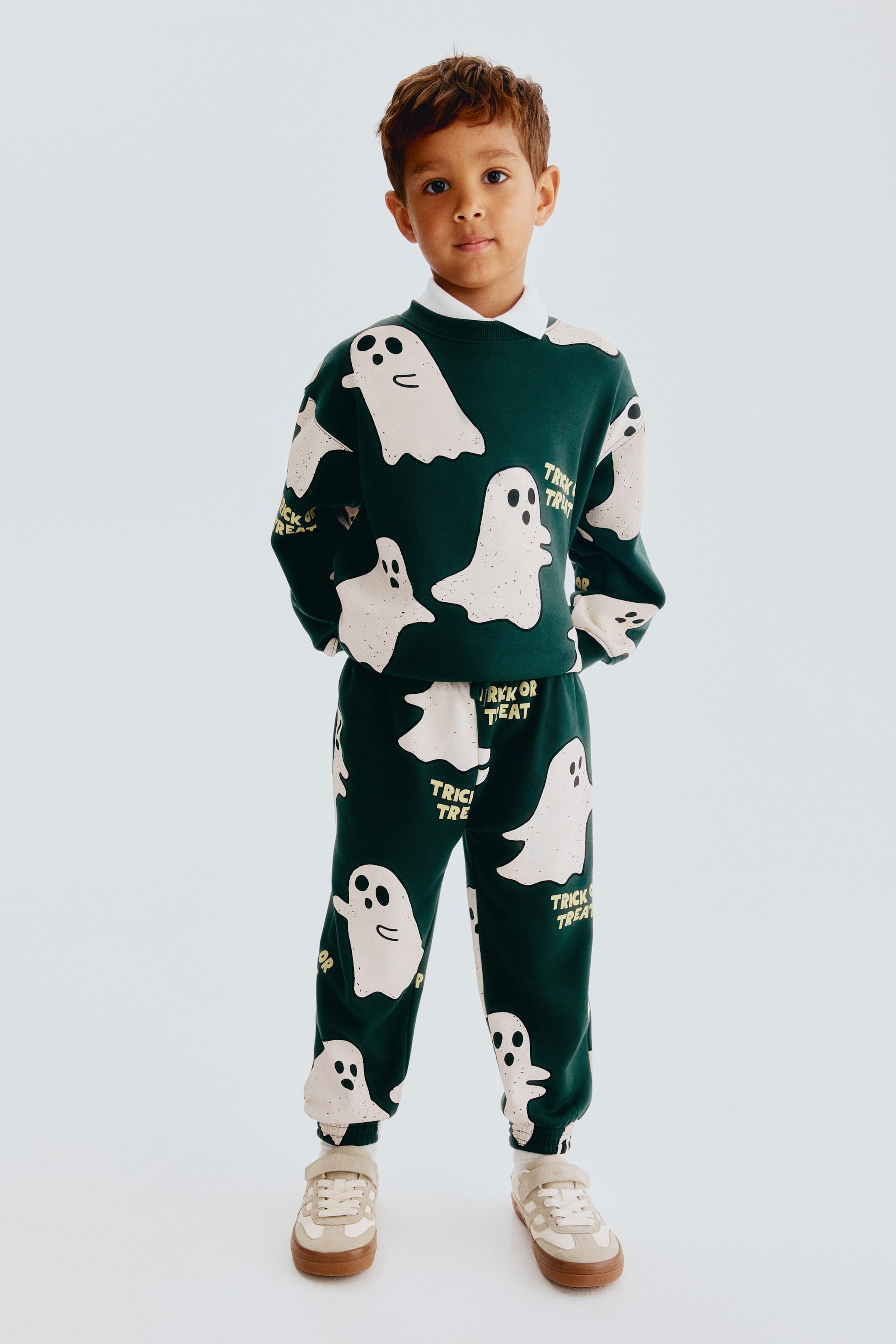 View larger image: A dark green two-piece loungewear set is shown from the front, featuring an allover print of light beige ghosts with black facial features and yellow "Trick or Treat" text. The crew-neck sweatshirt features ribbed cuffs and hem, with a white collared shirt peeking out from the neckline, and the matching sweatpants also feature ribbed ankle cuffs.