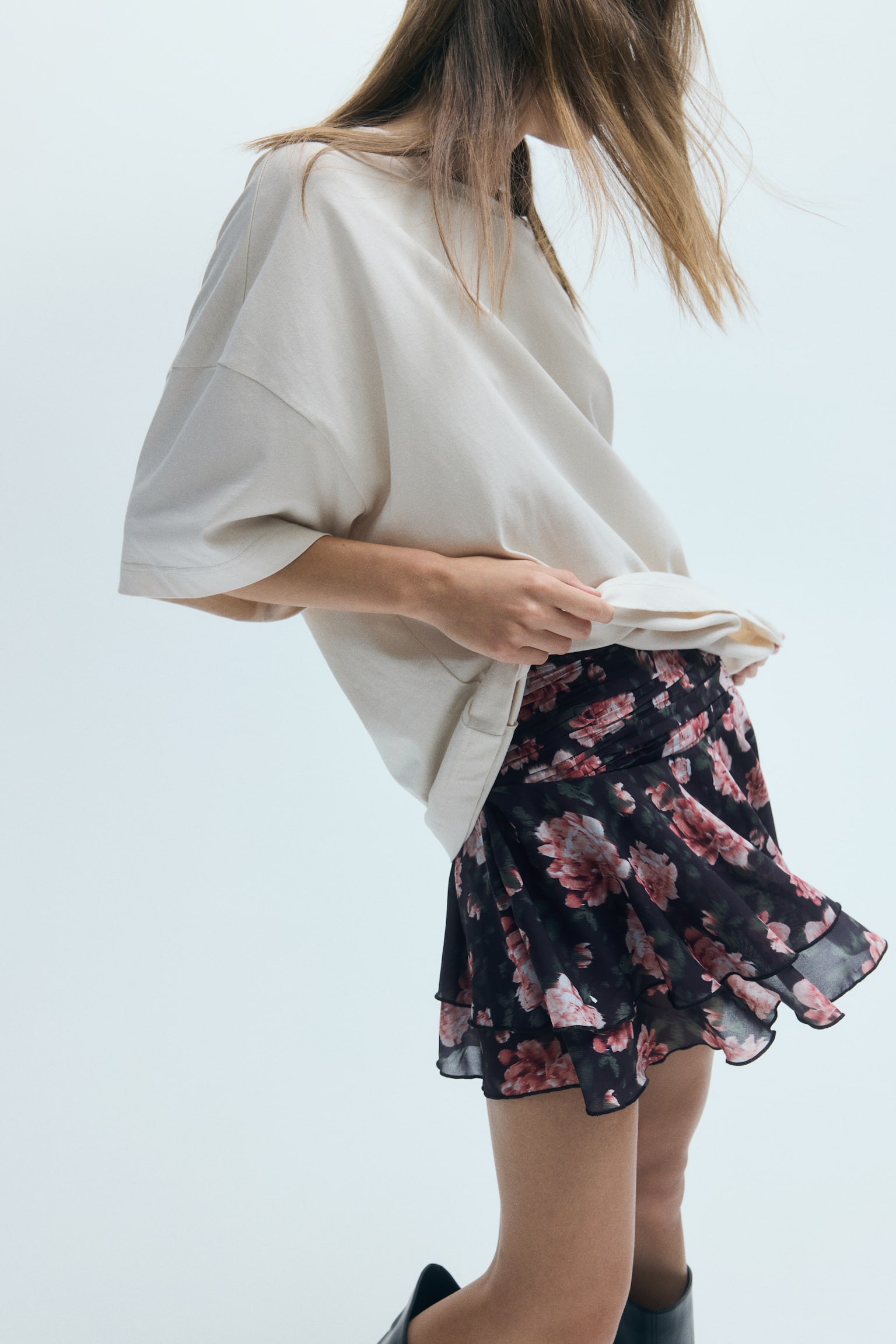Uma minissaia evasé com padrão floral preto e rosa, com folhos em camadas e transparência, está a ser usada pela modelo. A modelo está a levantar a bainha de uma t-shirt creme oversized para mostrar a cintura plissada da saia. - 1
