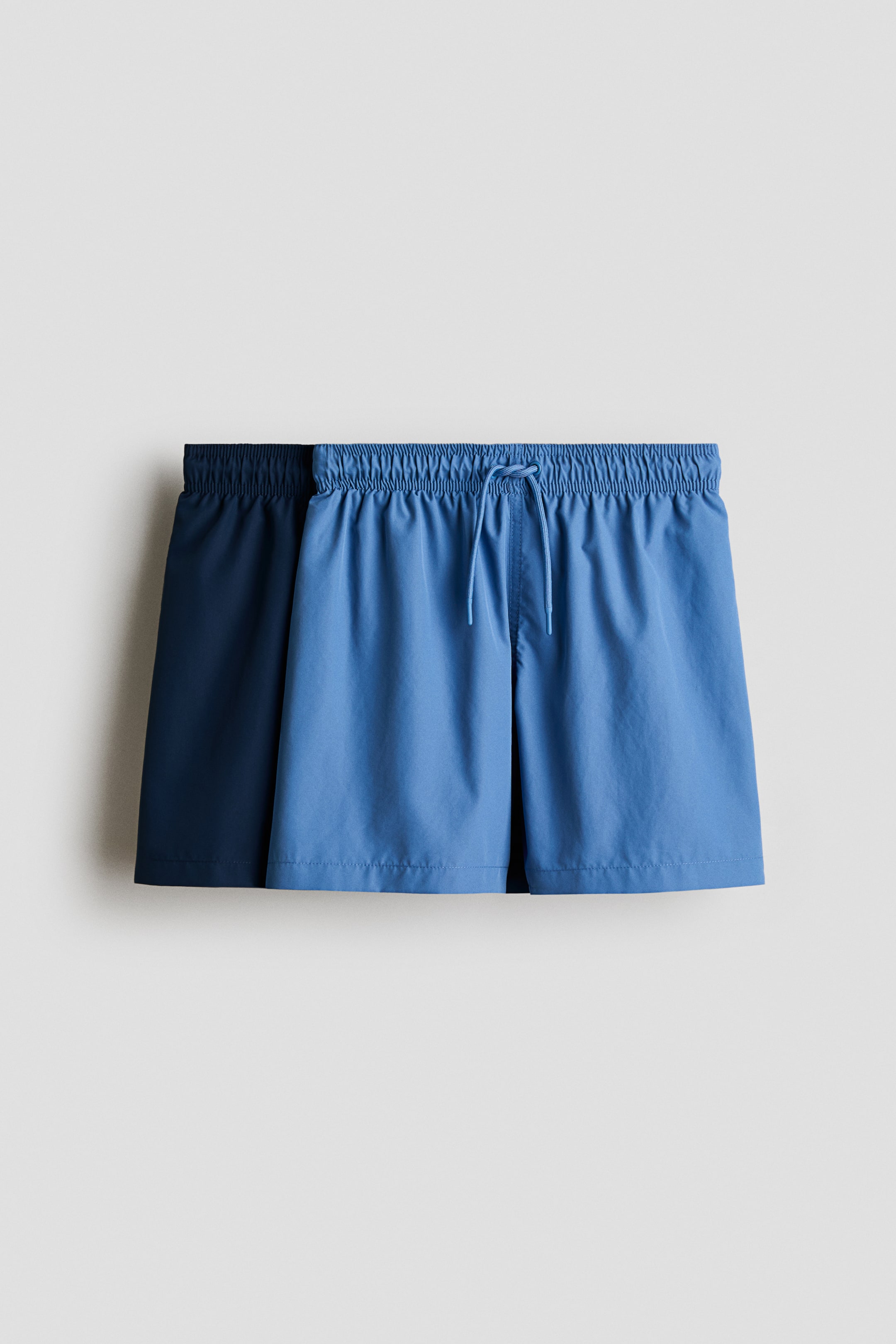 Vis større bilde: To par shorts vises forfra. Det mellomblå paret foran har en elastisk linning med synlig snor, mens et mørkeblått par delvis synlig bak det også viser en elastisk linning.