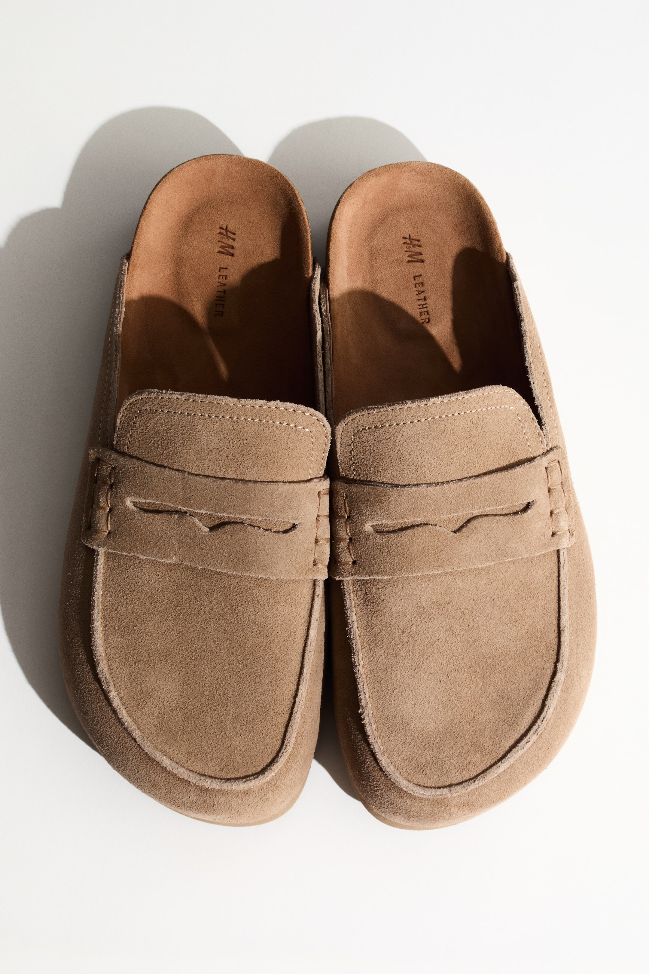 View larger image: A pair of light brown suede mule loafers is facing forward from a top-down perspective, featuring a penny loafer strap across the vamp, visible stitching around the toe box, and brown leather inner soles displaying "H&M LEATHER" branding.